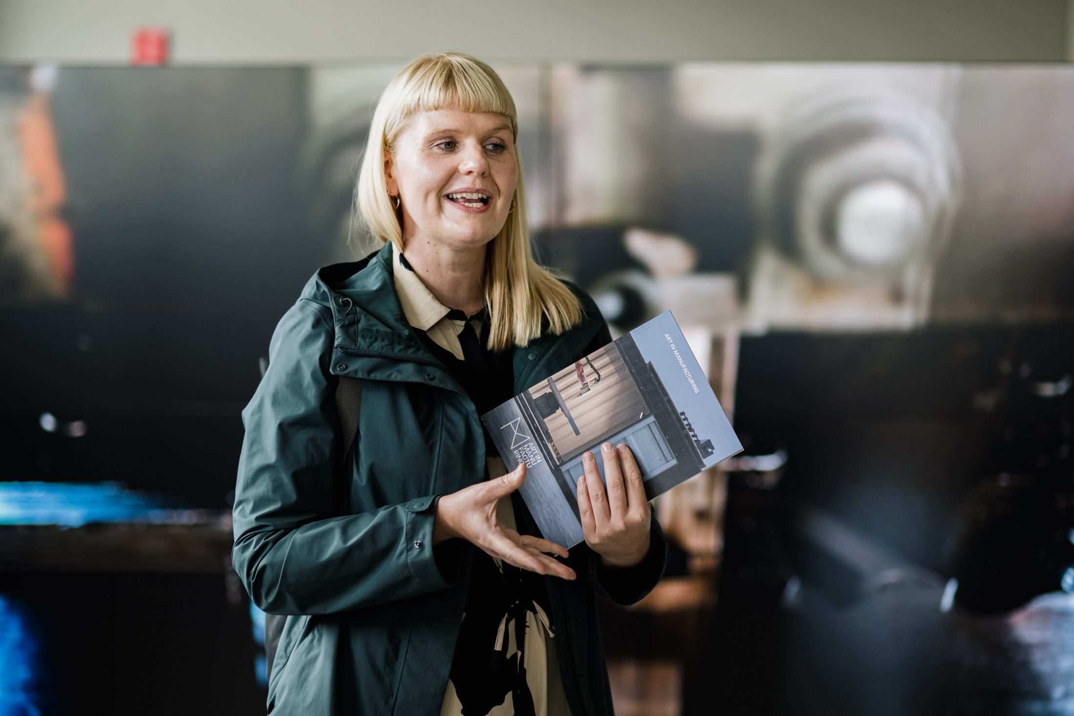 A woman with blonde hair and fringe, wearing a dark jacket, holds an open book and speaks in front of a blurred background.