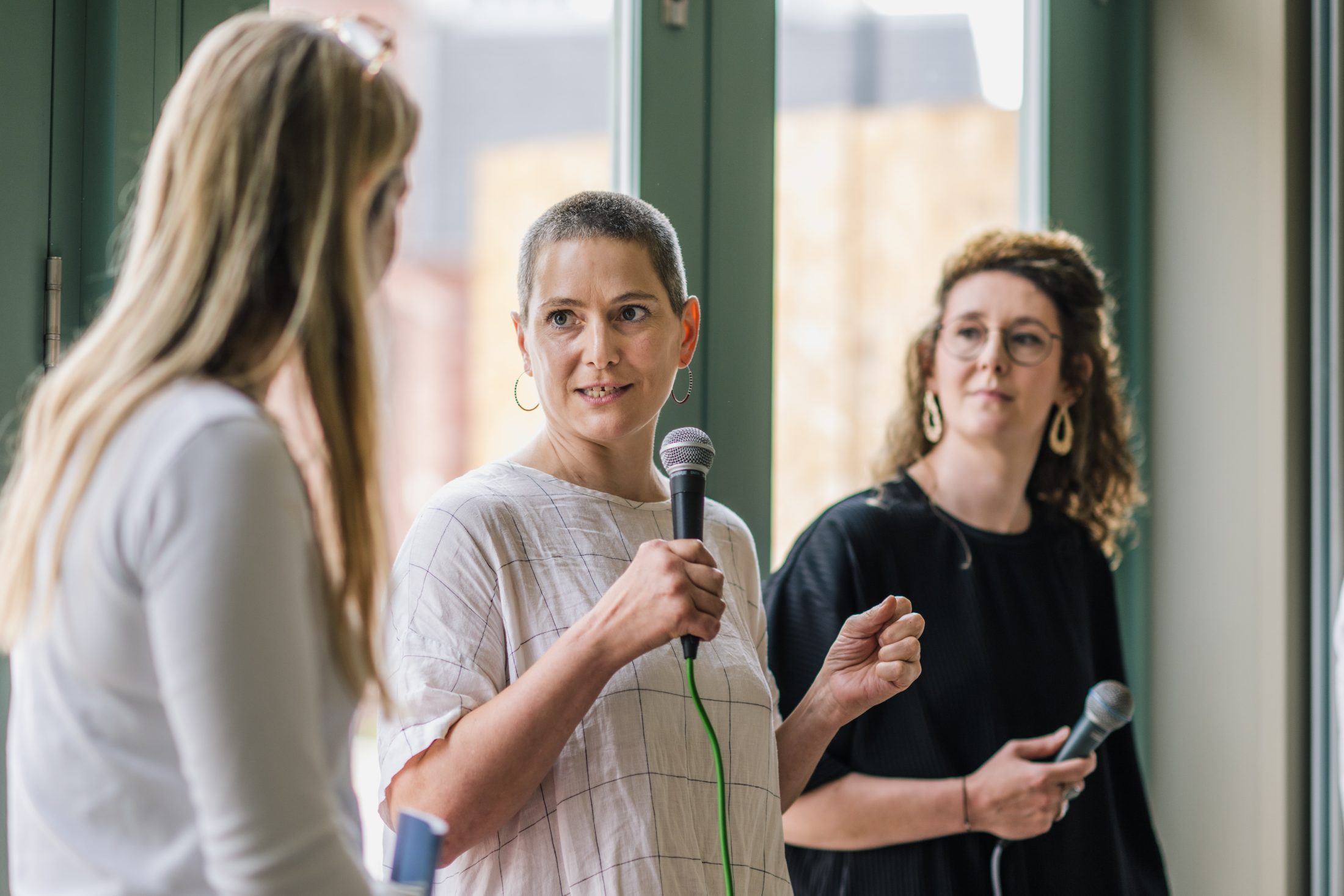 Three women stand indoors holding microphones. The woman in the centre is speaking, while the other two listen attentively. They are near large windows with natural light shining in.