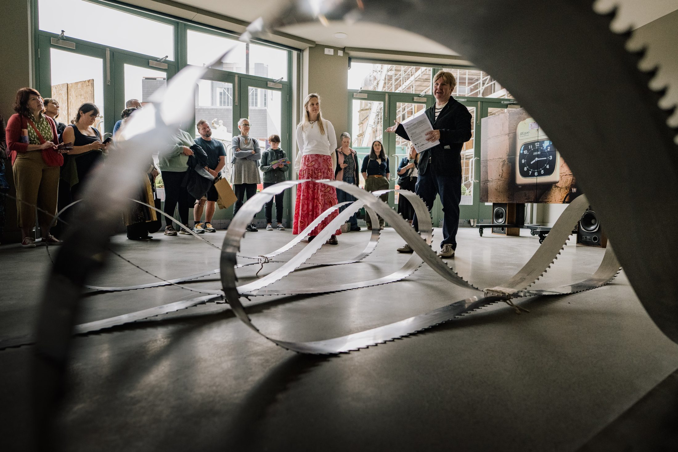 A group of people stands in a room with large windows, observing a modern, wavy metal sculpture on the floor. Two people at the front appear to be presenting or speaking to the audience.