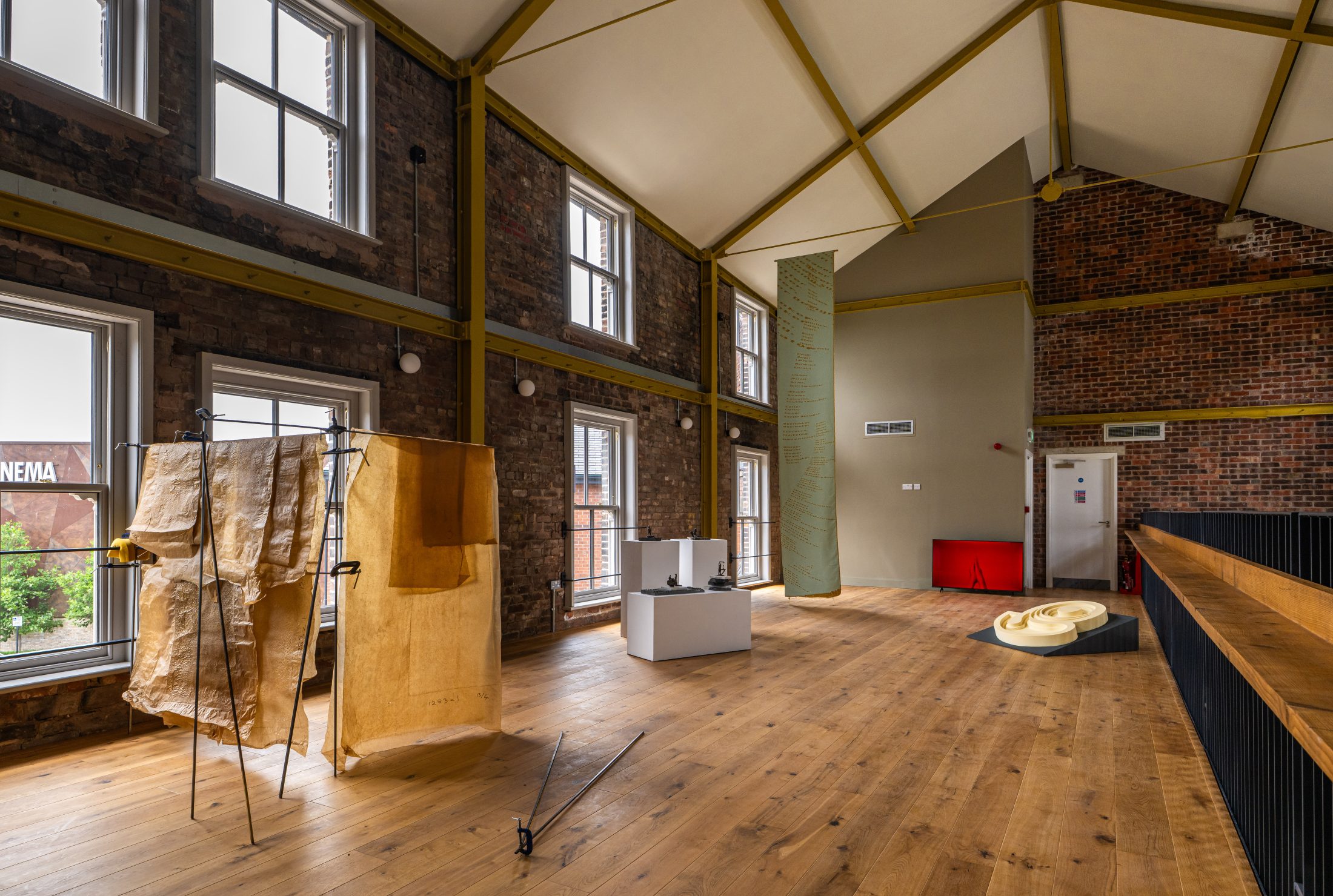 A spacious art gallery with tall ceilings, large windows, exposed brick walls, wooden floors, and minimal artworks displayed, including draped fabric, white plinths, and a yellow abstract sculpture on the floor.