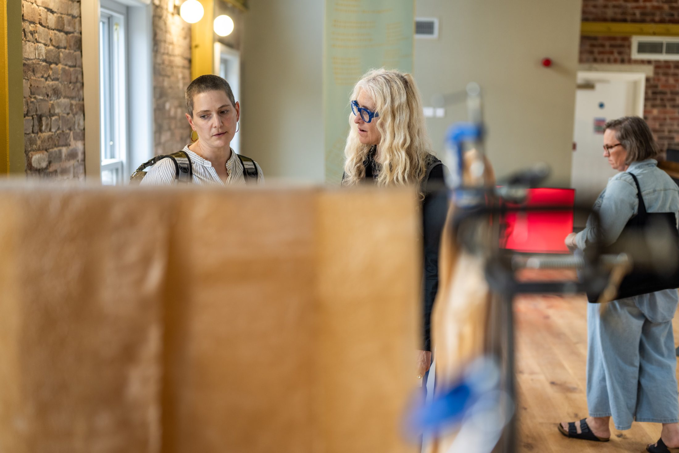 Two women closely observe an art piece in a gallery whilst another woman stands in the background, all in a well-lit room with brick walls and wooden floors.