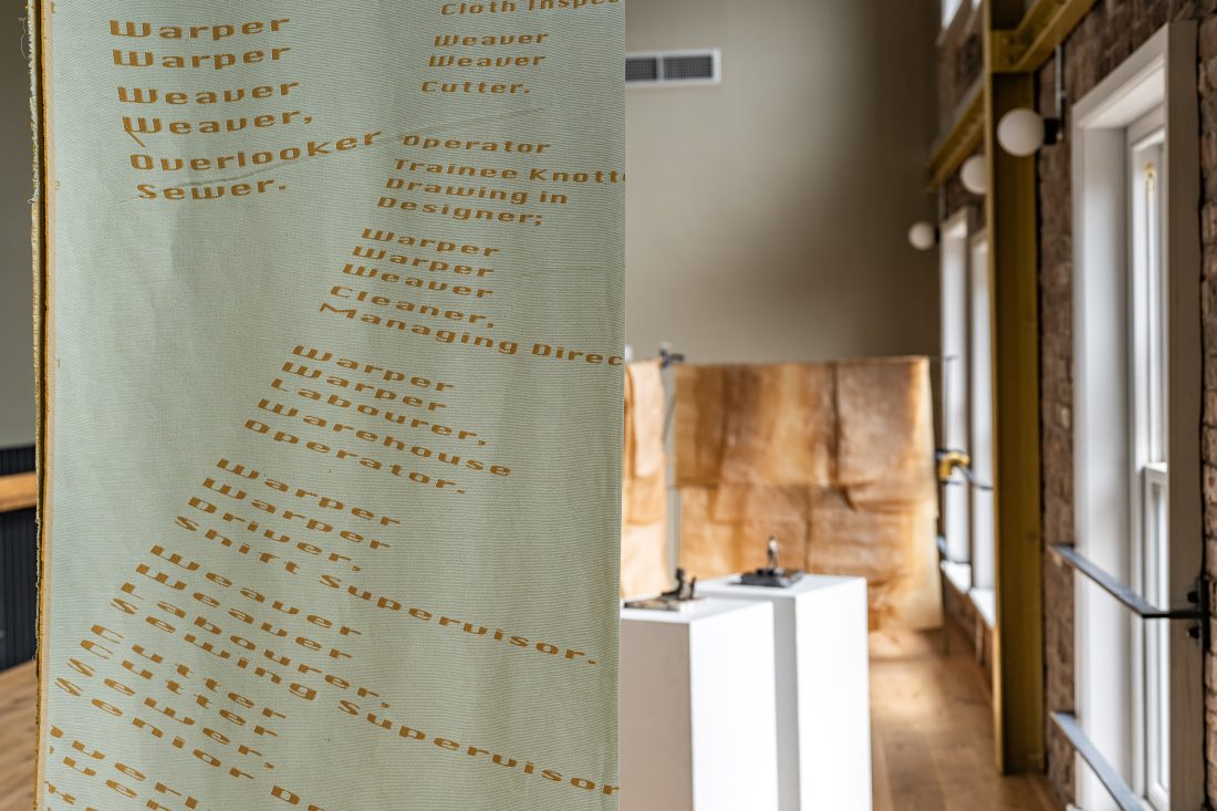 A fabric panel lists textile job titles like warper, weaver, and cutter in yellow text. The panel hangs in a modern, airy room with wooden floors, tall windows, and minimalistic white display plinths.