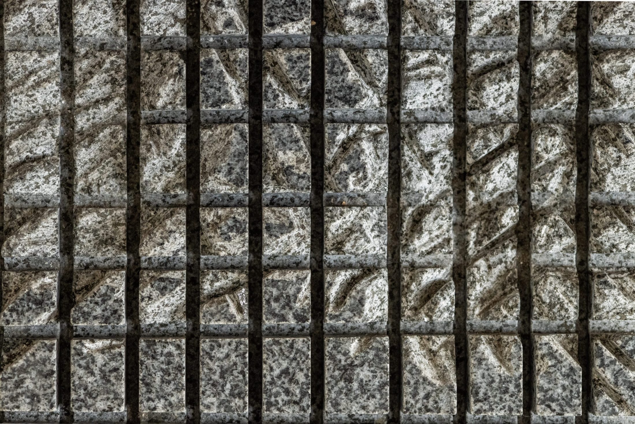 Close-up of a textured stone wall with a grid of rectangular sections, each separated by dark vertical and horizontal grooves, creating a chequered pattern with varying shades of grey and rough surface details.
