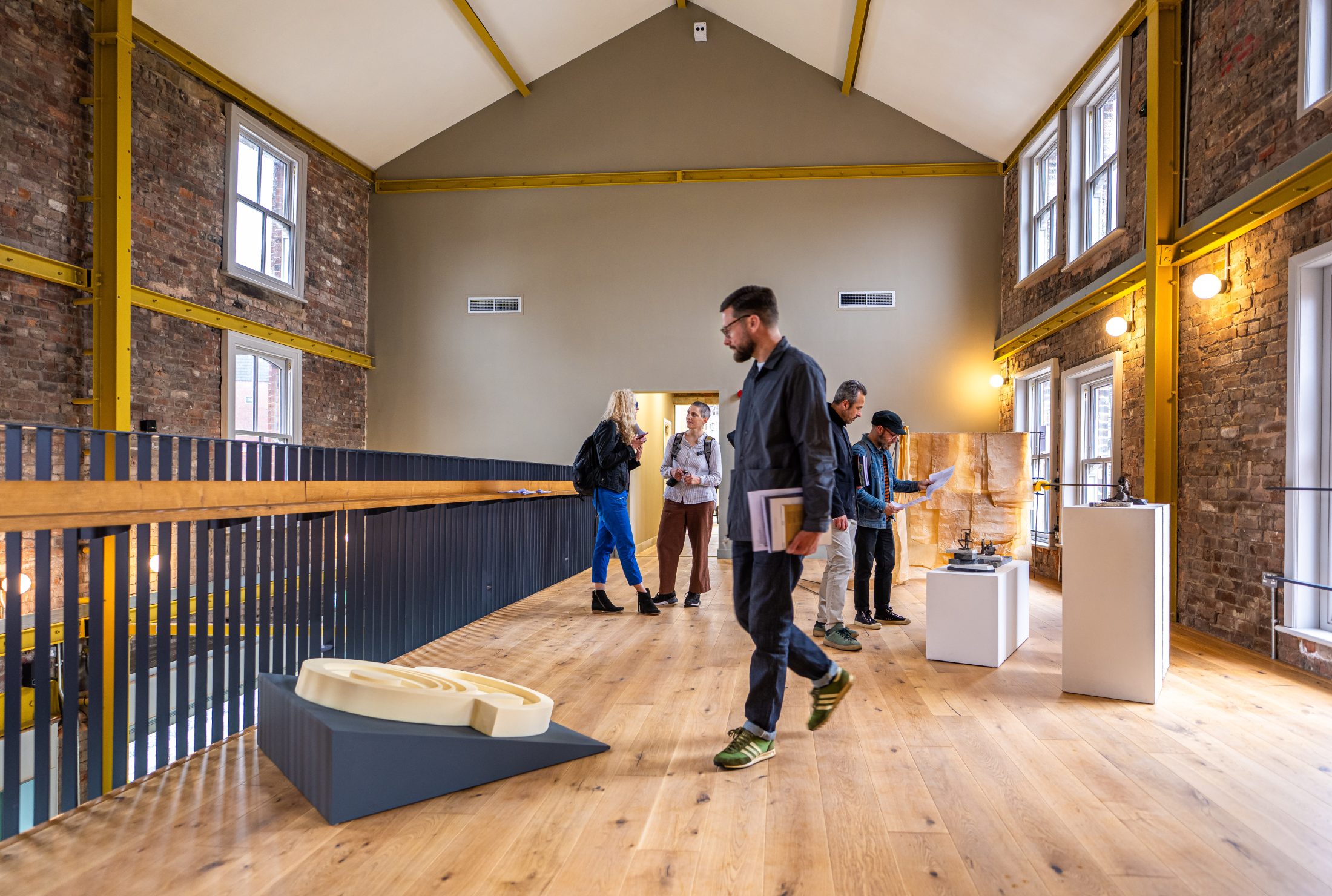 A group of people views modern art sculptures displayed on plinths and the floor inside a spacious, high-ceilinged gallery with exposed brick walls and large windows.