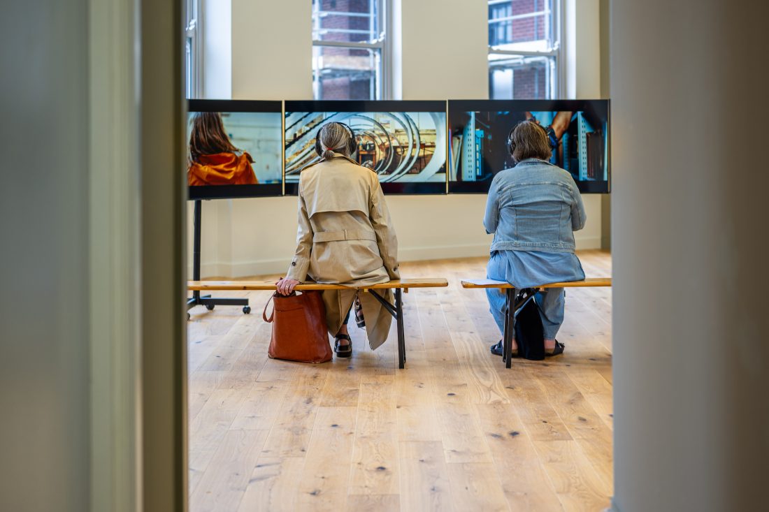 Two people sit on a bench in front of a row of large screens displaying images in a modern, bright room with wooden floors and large windows.