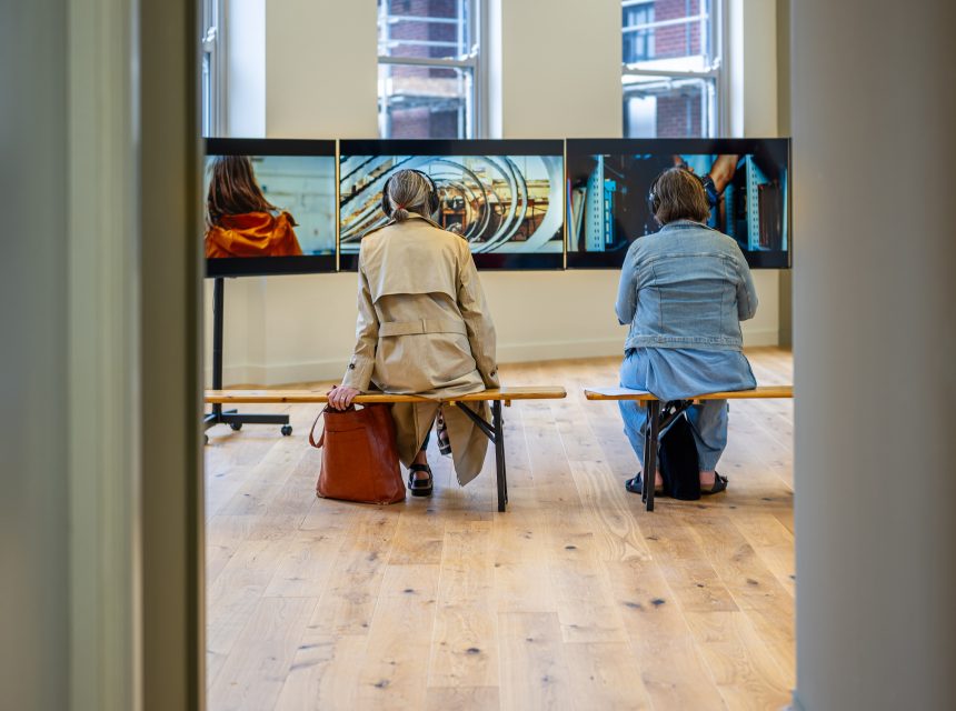 Two people sit on a bench in front of a row of large screens displaying images in a modern, bright room with wooden floors and large windows.