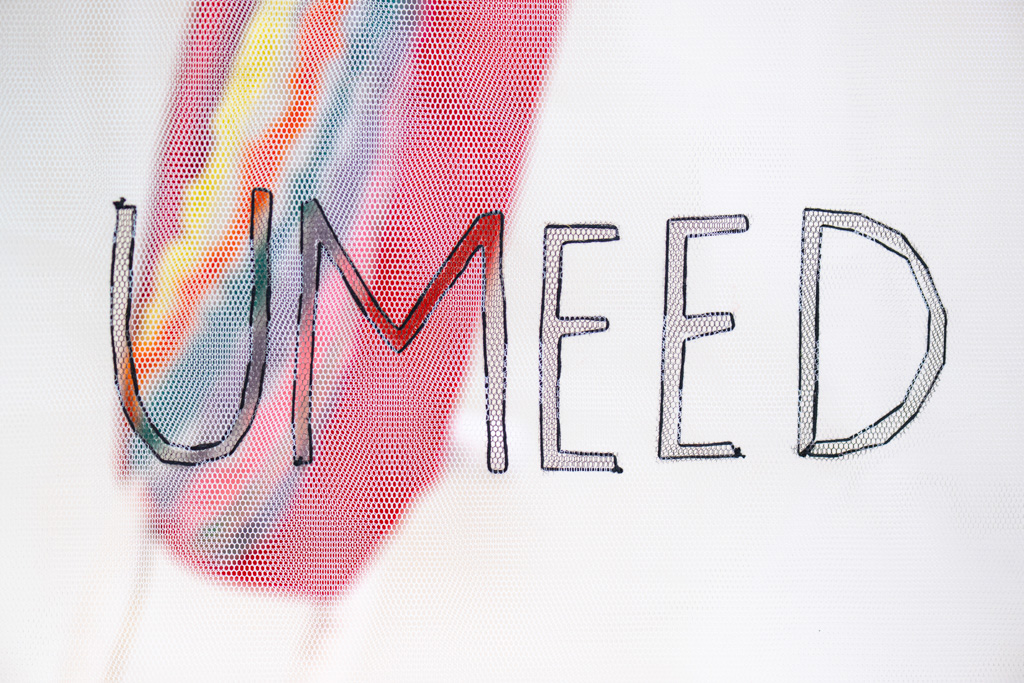 The word UMEED is handwritten in large, outlined letters on a white background with a blurred, multicoloured streak running diagonally behind the text.