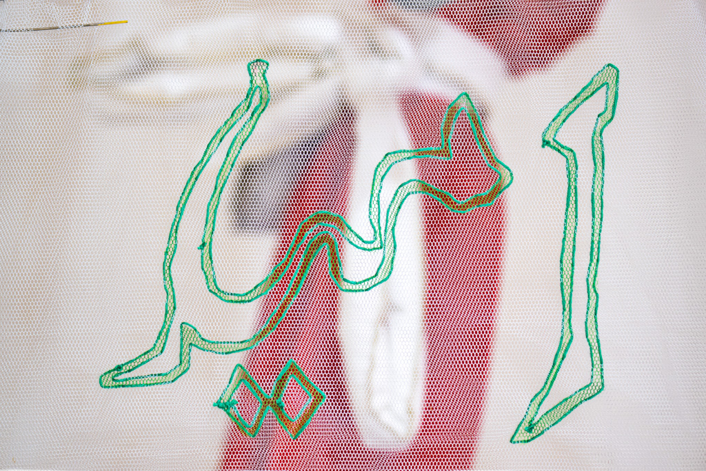 Green Arabic calligraphy embroidered on white mesh fabric, with a blurred background showing red and white shapes.