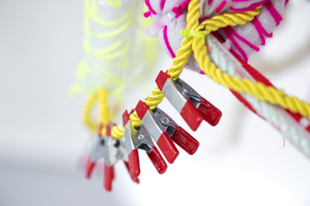 Close-up of red pegs clipped to a yellow washing line, with colourful, fuzzy materials in the background, including pink and yellow patterns, against a white backdrop.