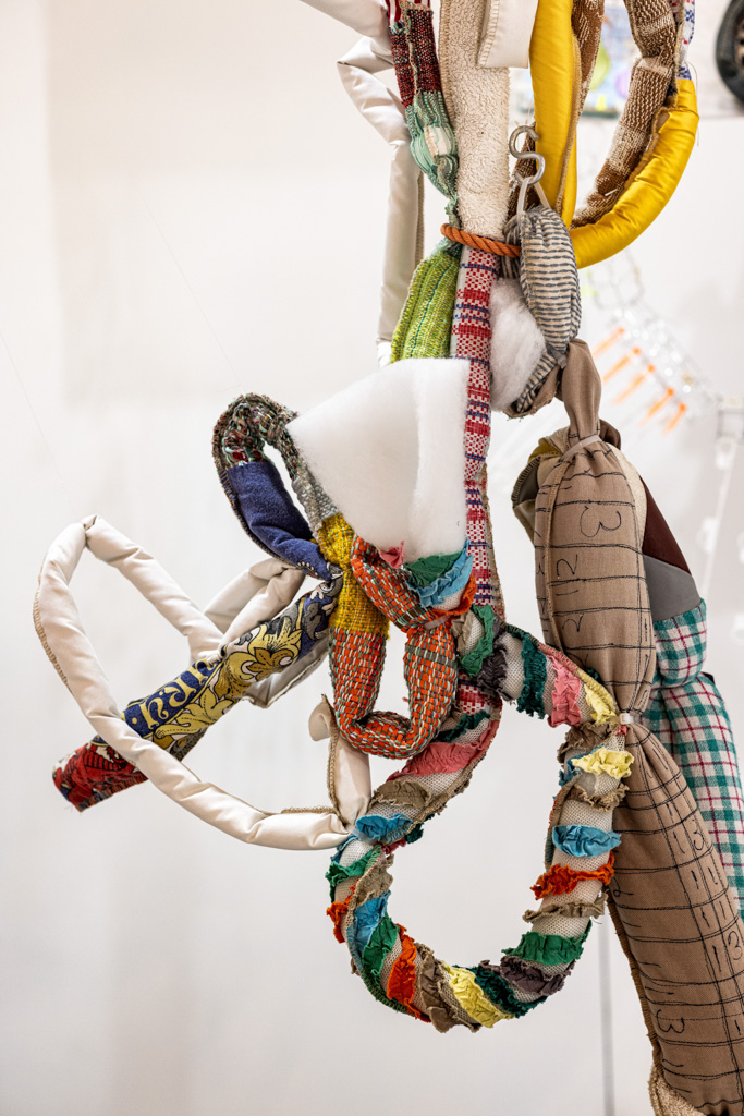 A close-up of a hanging sculpture made from various colourful, textured fabrics, cords, and padded materials twisted and knotted together in loops and abstract shapes.
