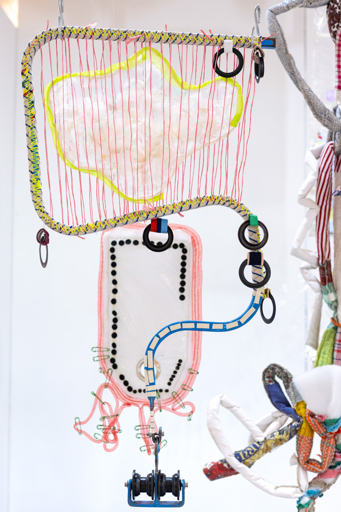 Colourful abstract art sculpture featuring two rectangular frames adorned with wires, rings, and fabric. The upper frame has red vertical strings and a cloud-like shape, while the lower one features loops, black dots, and a blue, tube-like attachment.