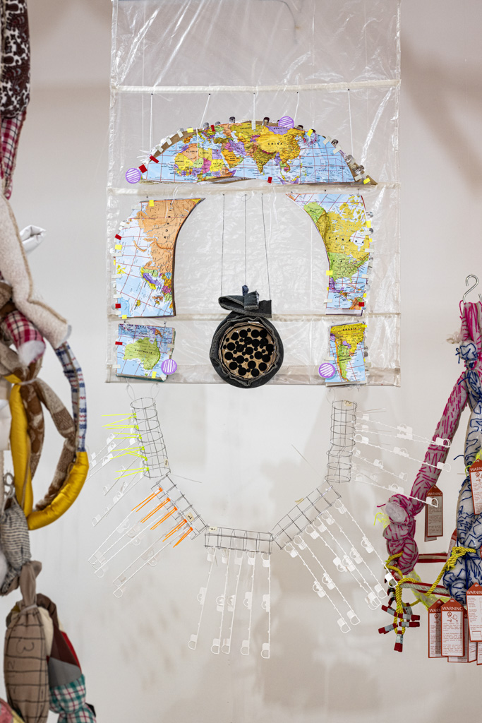 A hanging art piece made from maps, plastic, and metal clips, arranged in an arch shape with a round, black object in the centre. Colourful cloth-wrapped objects hang on either side.