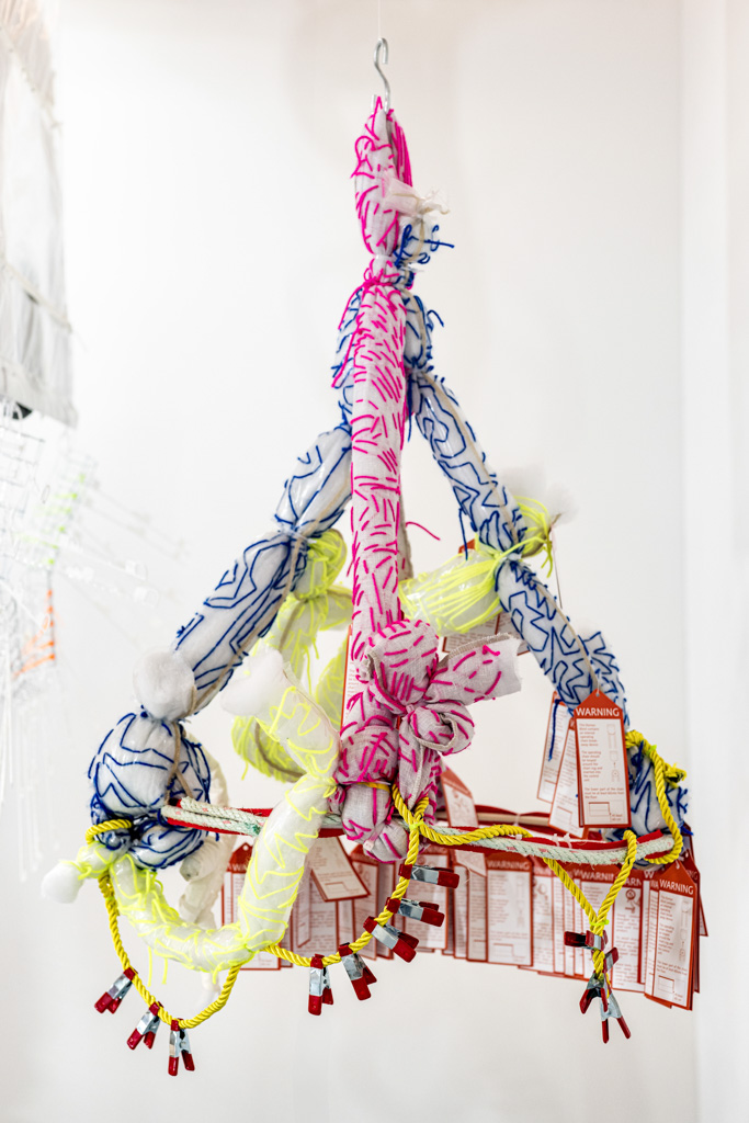 A colourful hanging sculpture made of patterned fabric tubes, yellow rope, white pom-poms, red tags, and red clips, suspended from the ceiling against a white background.