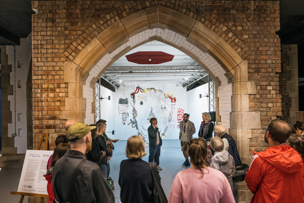 A group of people listen to two speakers inside a brick-arched gallery space, facing a white wall with colourful, abstract art and decorations. An information board is visible on the left.