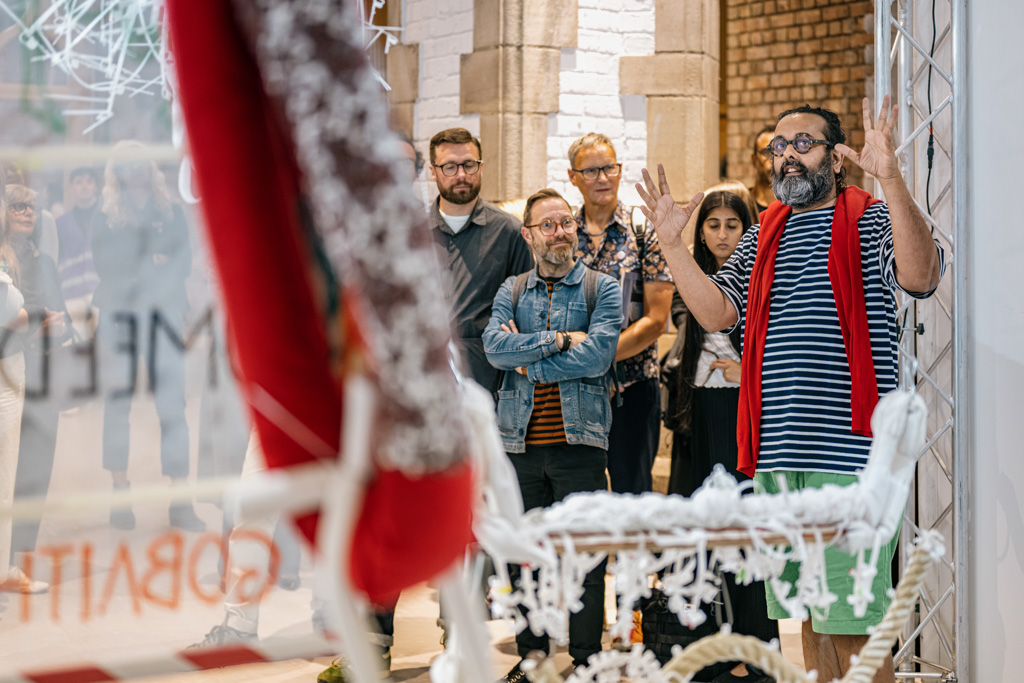 A man with glasses and a red scarf speaks animatedly to a group of attentive people in an art gallery. Crochet artwork and installation pieces are visible in the foreground.