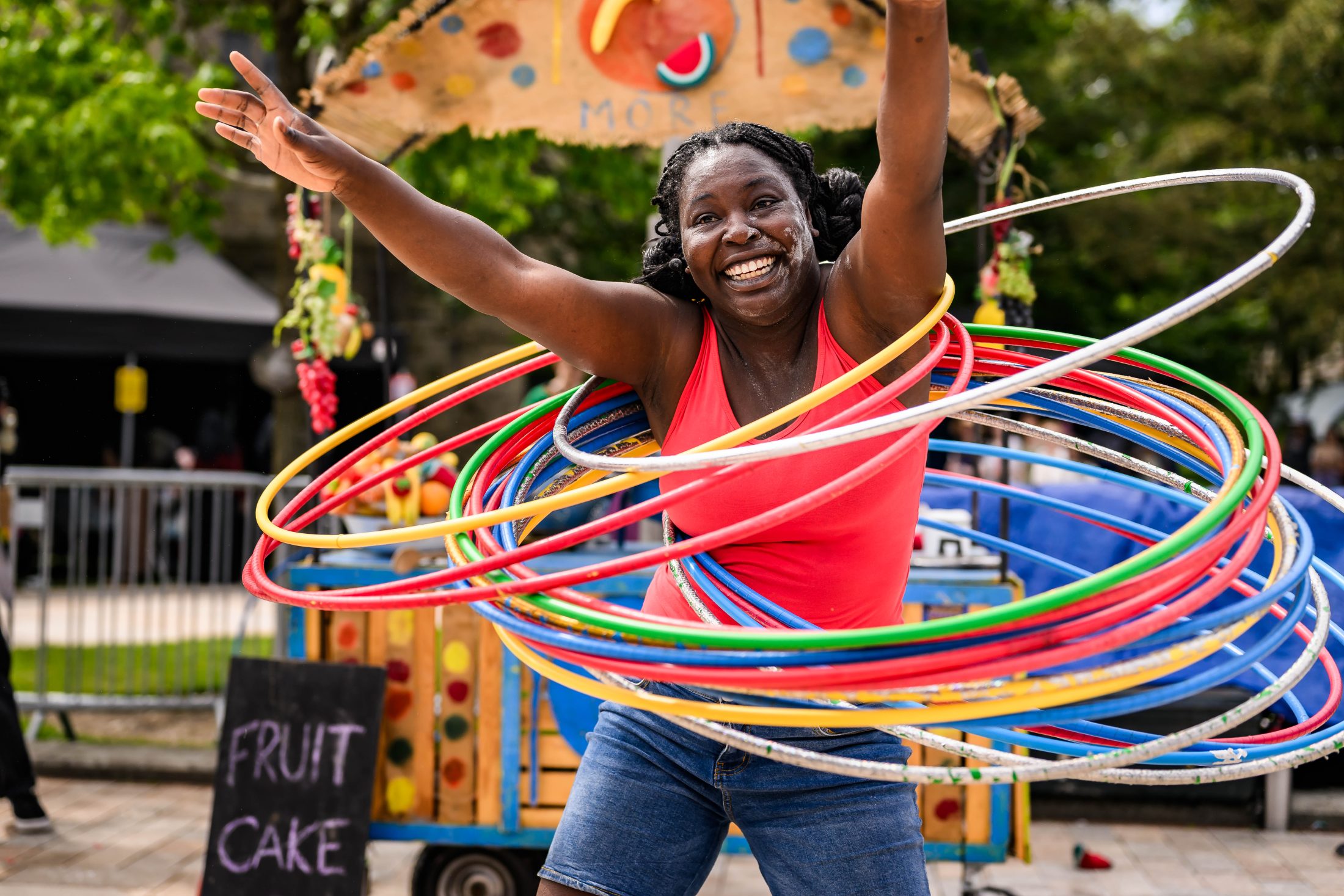 A woman in a bright pink vest smiles joyfully whilst spinning multiple colourful hula hoops around her waist, arms raised, at an outdoor event with a “Fruit Cake” sign in the background.