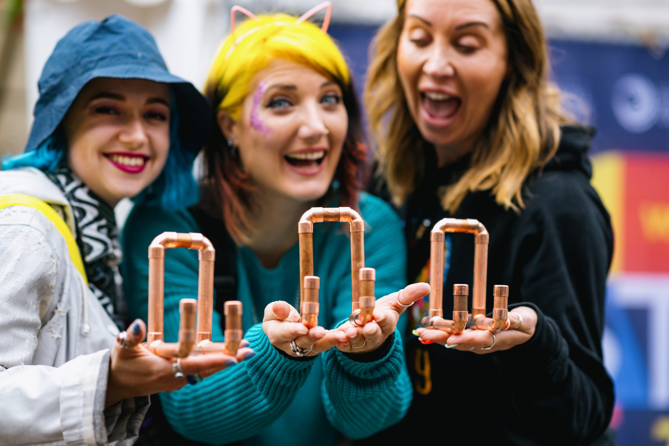 Three smiling women hold up copper pipes assembled into geometric shapes, posing happily together. One woman wears a blue hat, another has blonde hair and cat ears, and the third is in a black hoodie.
