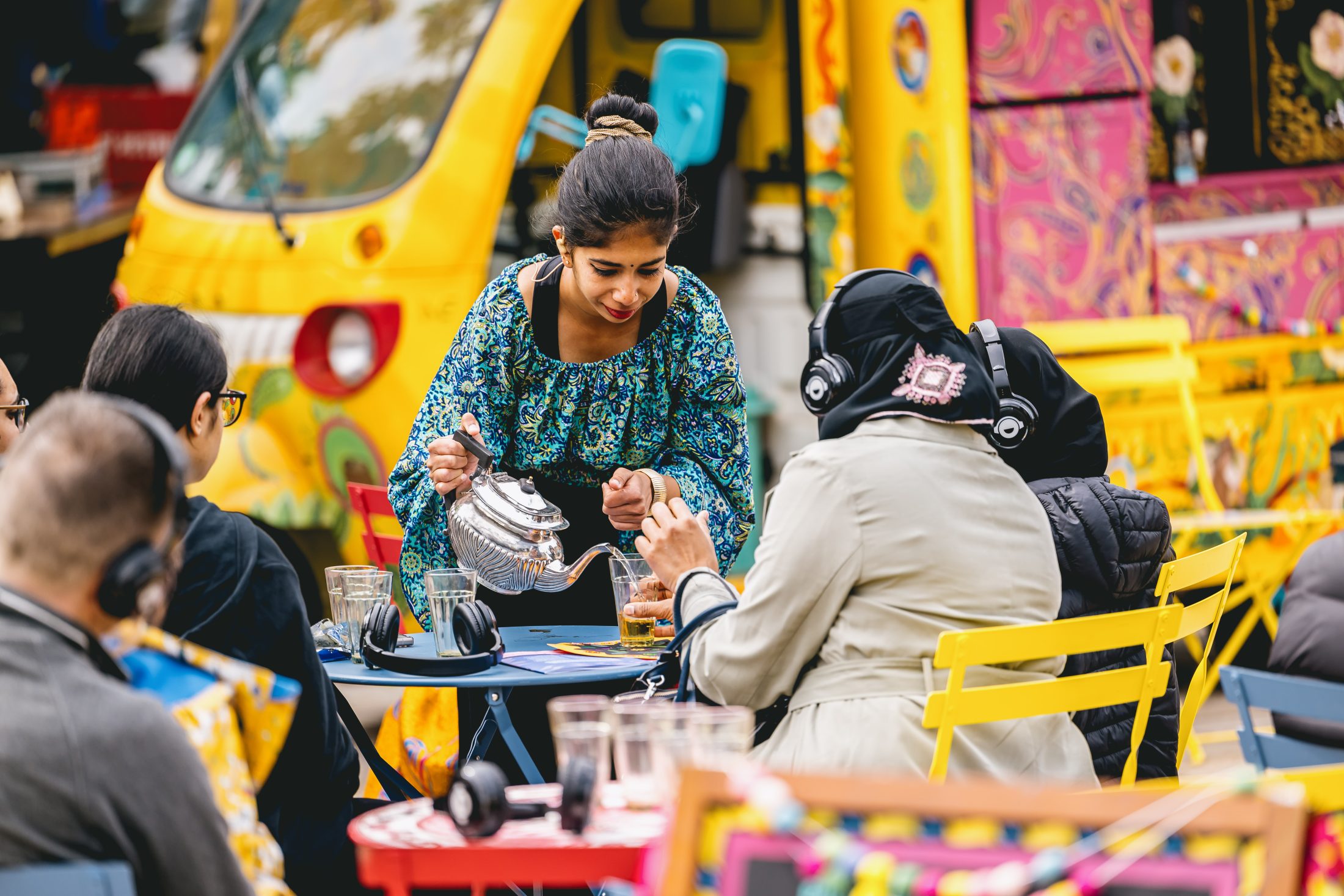 A woman in a blue-patterned dress pours tea from a silver teapot for people sitting at colourful outdoor tables, with vibrant yellow and patterned decor in the background.