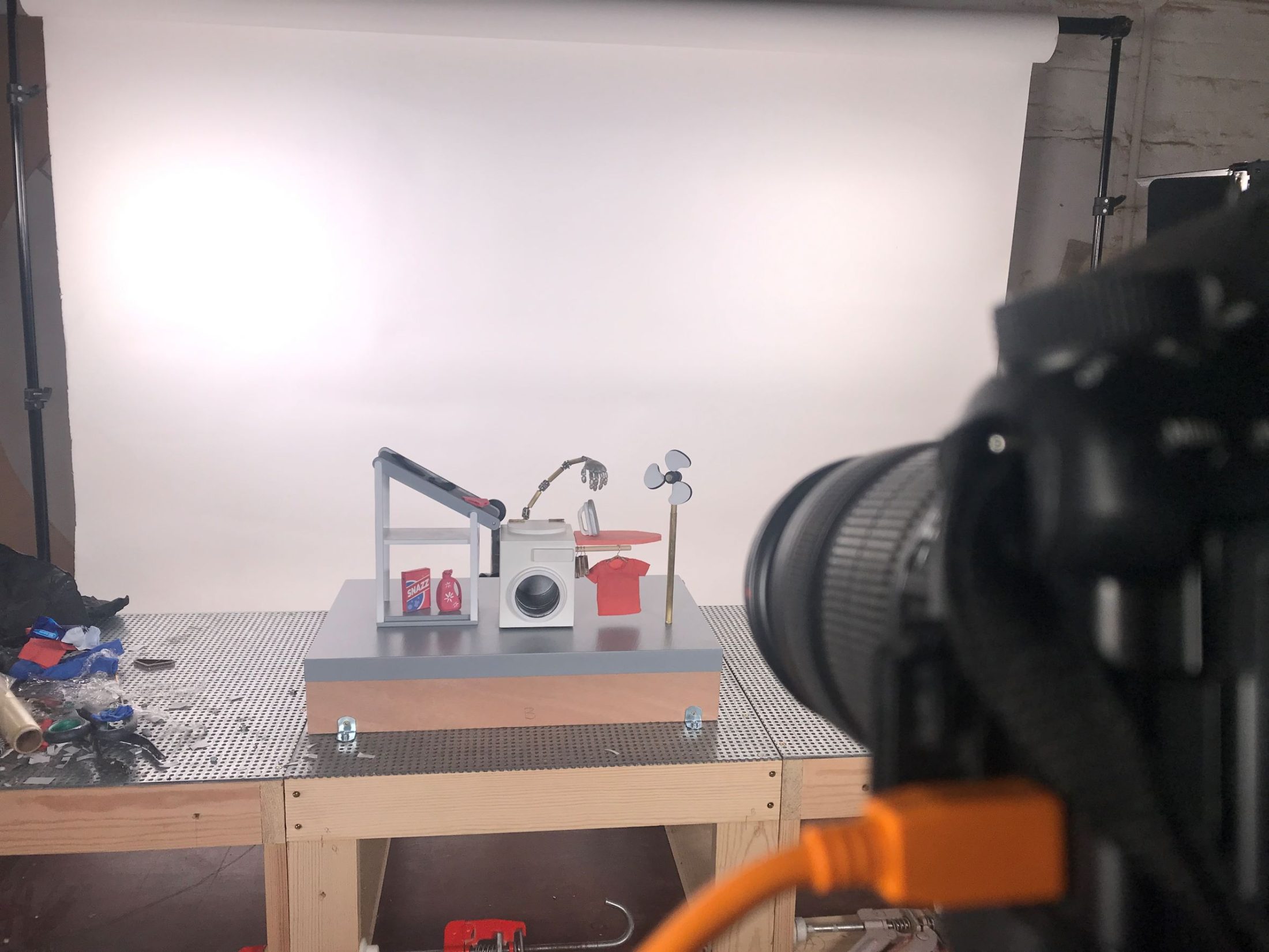 A camera films a small Rube Goldberg machine model on a table, set against a white backdrop in a photography studio. Various props, tools, and cables are visible around the set-up.
