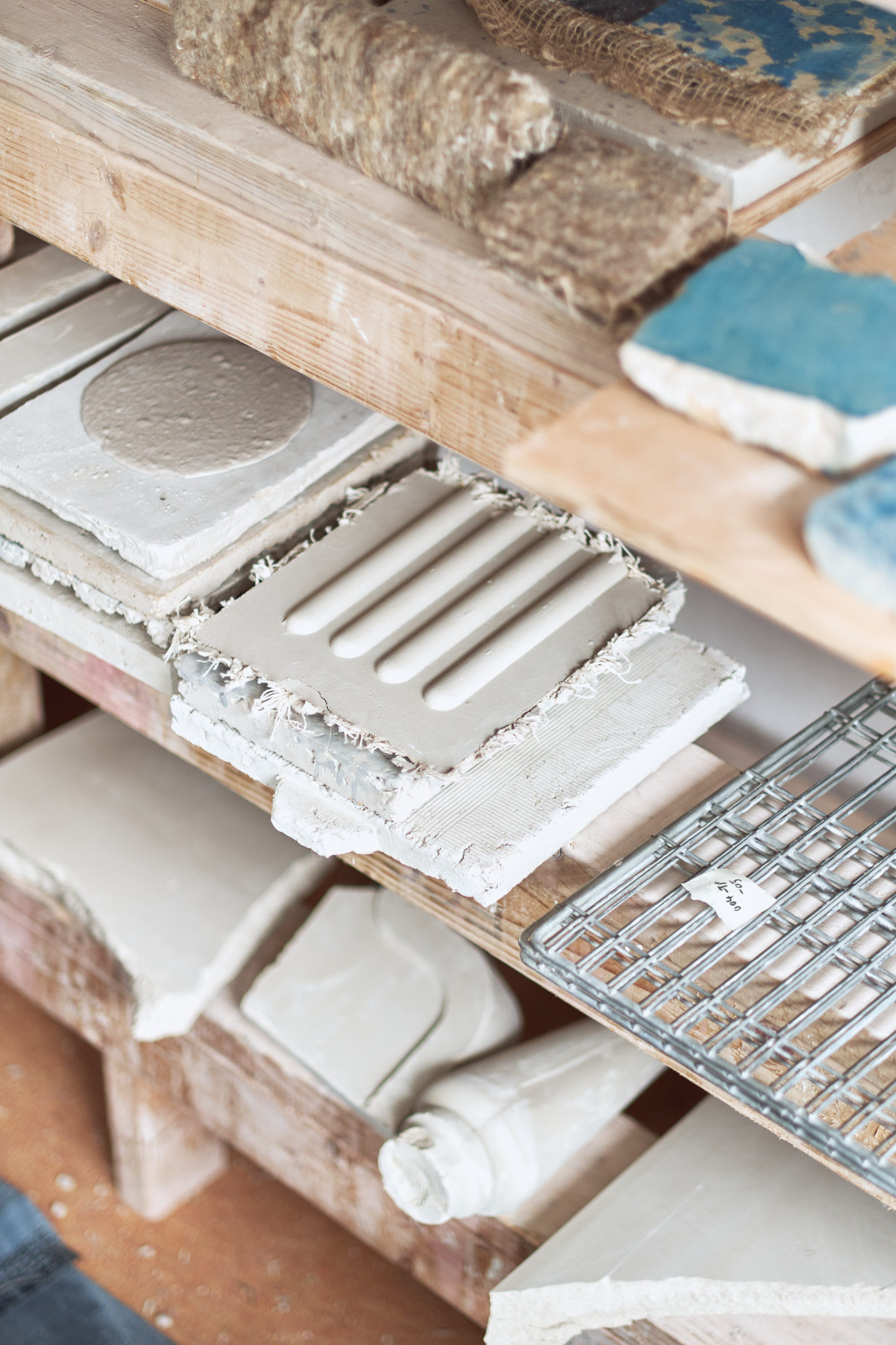 Shelf holding plaster moulds, textured clay tiles, a metal grid, and various art material samples, with some rough, unfinished edges and natural, earthy tones.