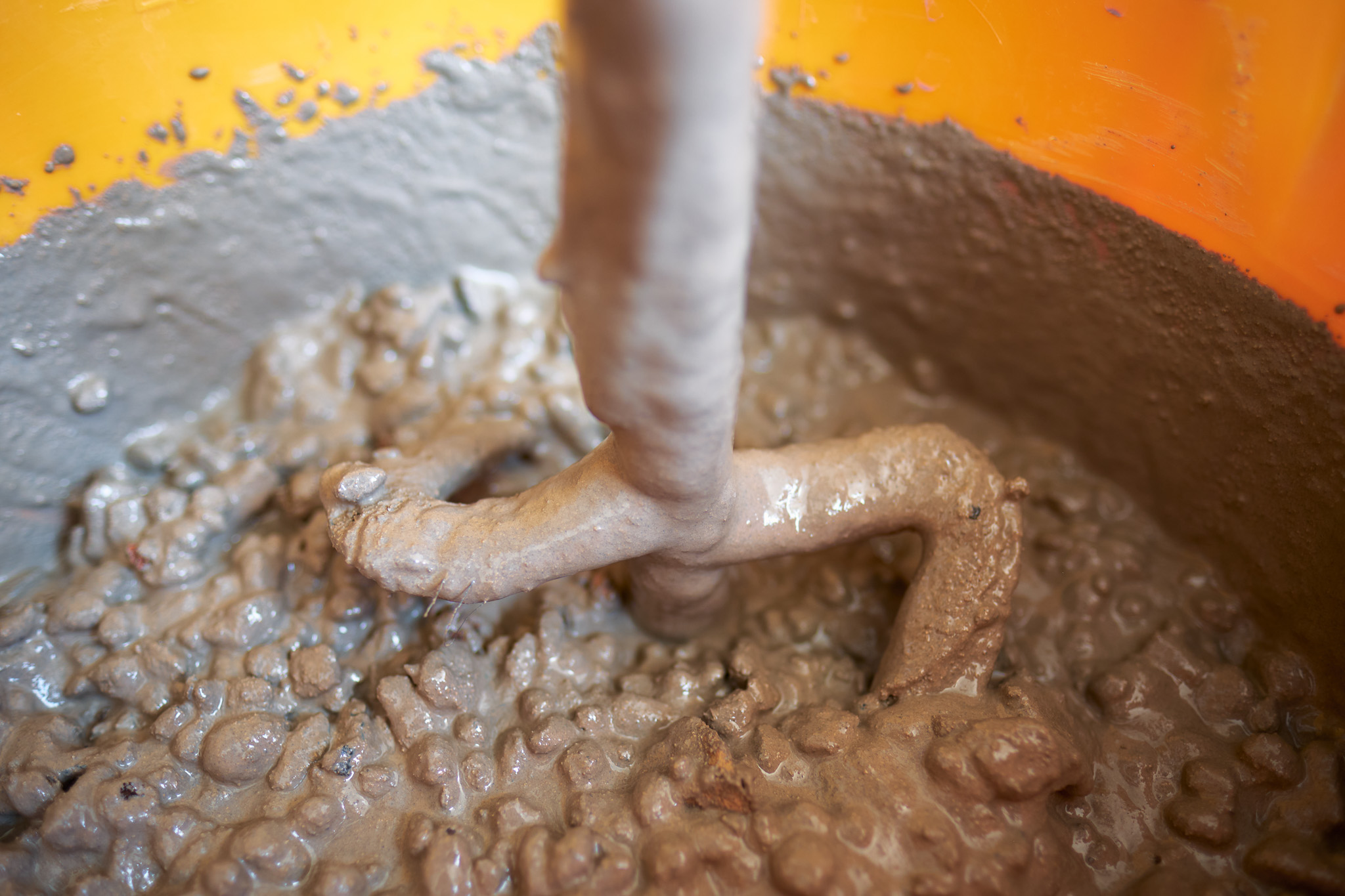 A close-up of a mixer stirring thick, wet concrete in an orange container, with bubbles and lumps visible in the grey mixture.