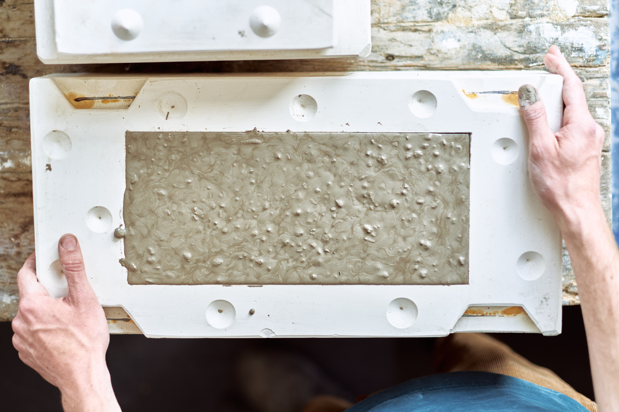 A person holds a rectangular white mould filled with wet, textured concrete or clay, likely for casting, on a worn wooden work surface.