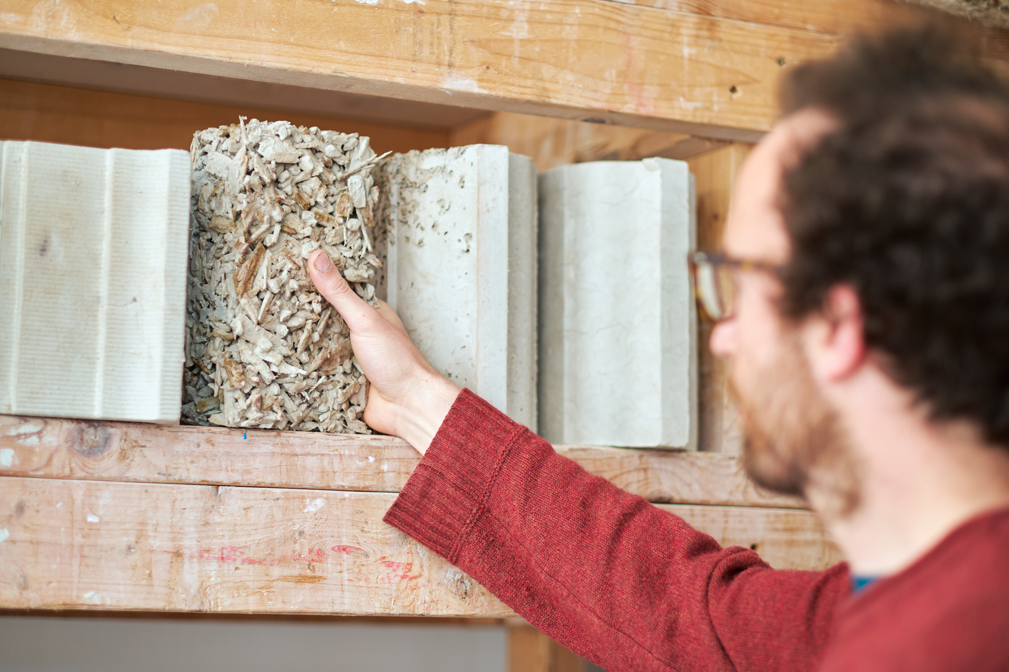 Lewis Jones in a red jumper reaches for a textured, fibrous building material sample on a wooden shelf, surrounded by other rectangular samples of various smooth materials.