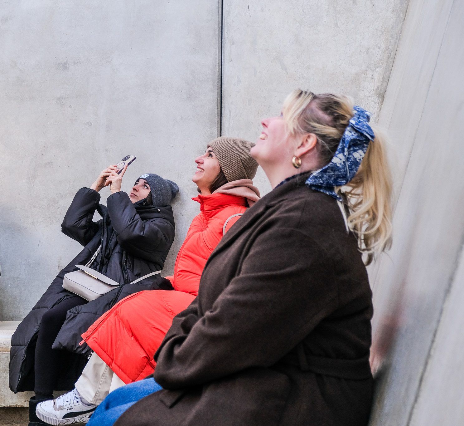 Three women in winter coats sit on a concrete bench, looking and smiling upward. One woman in a grey hat takes a photo with her mobile, while the others appear to be enjoying the view.
