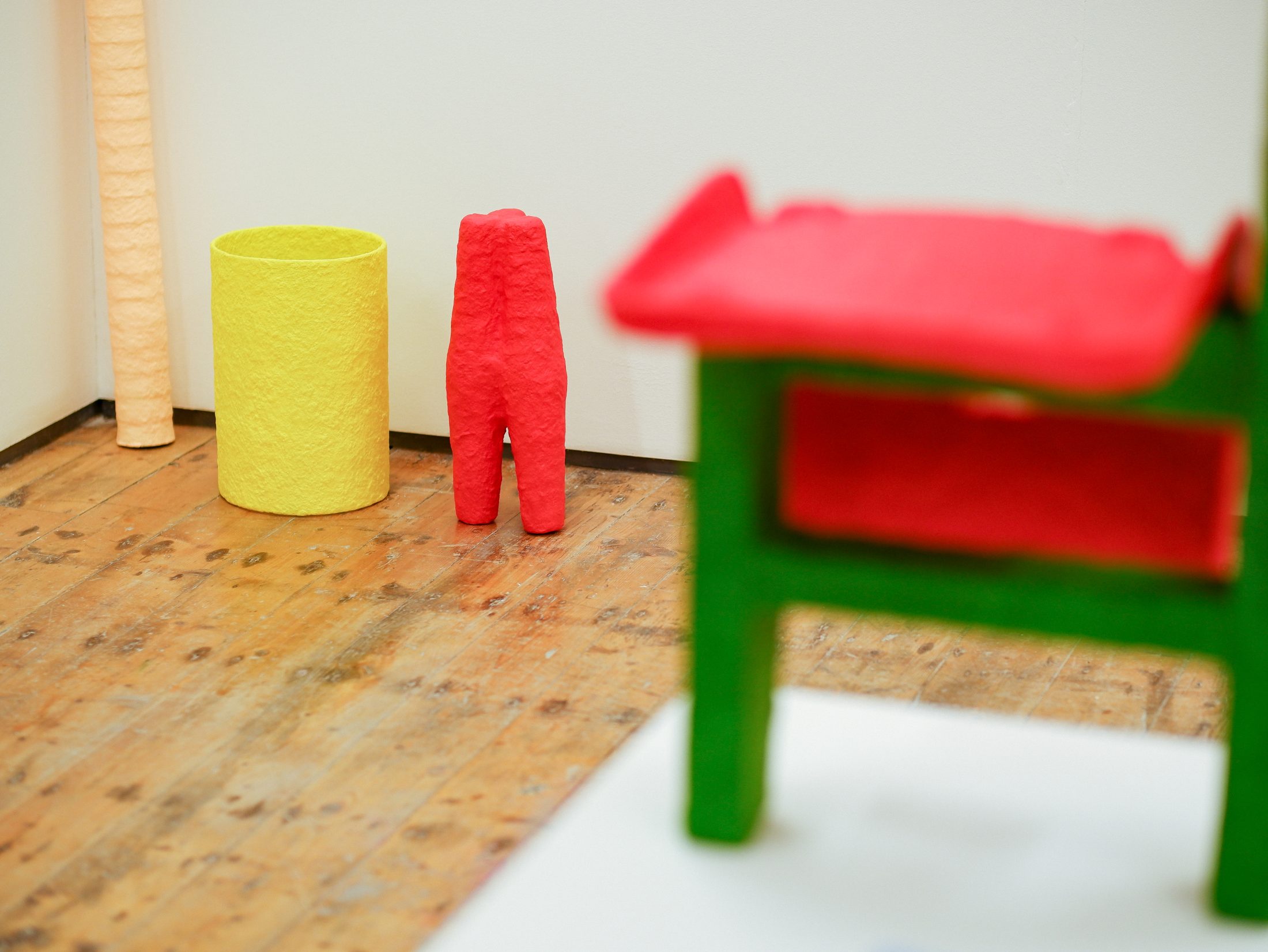 Vibrant abstract sculpture installation in a gallery. Features a red figure and yellow cylinder on a wooden floor, with a blurred green and red chair.