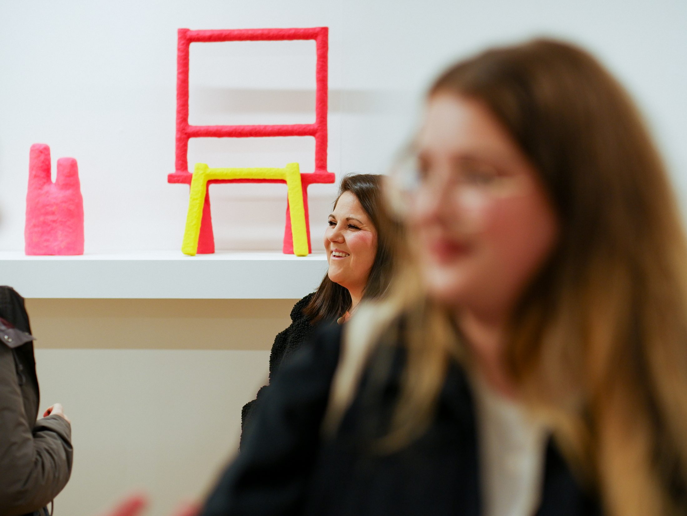 Smiling woman in a gallery with pink and yellow abstract art on a white shelf. Foreground blurred.