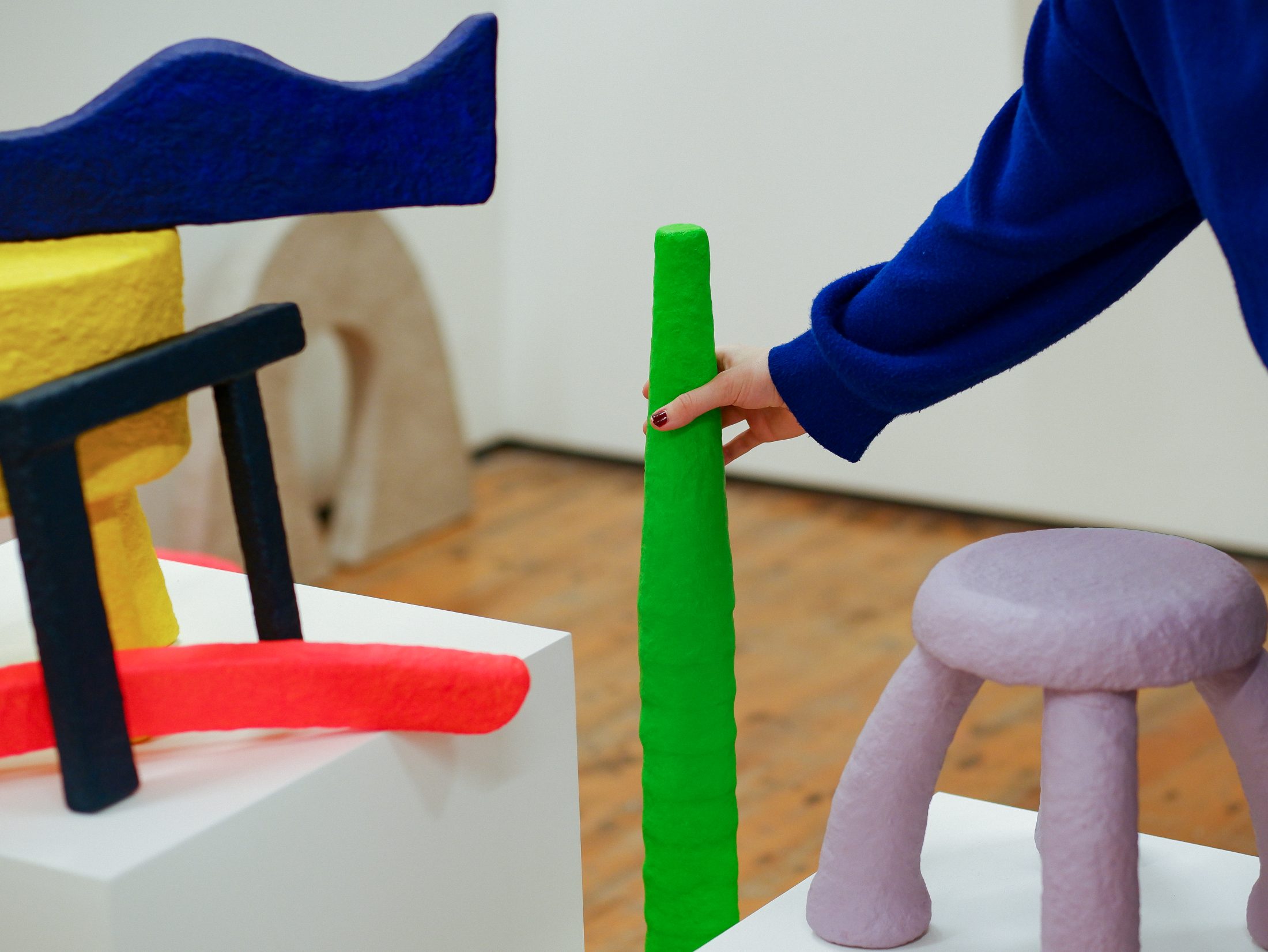 A person in a blue sleeve touches a tall, textured green sculpture among colourful, abstract objects on white plinths in an art gallery with wooden floors.