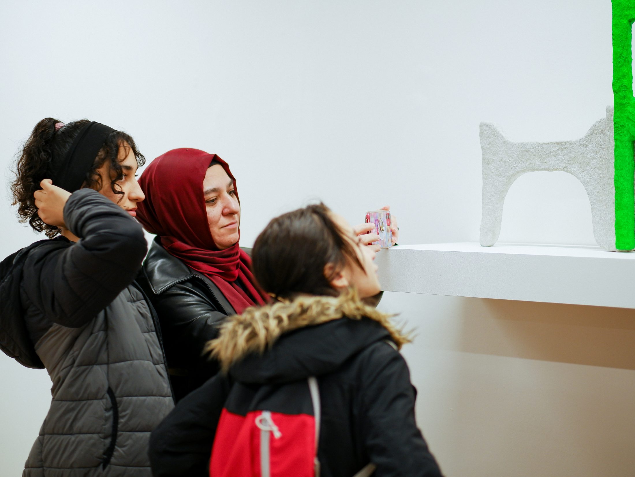 Three people observe abstract sculptures on a white shelf, with one taking a photo on their phone.