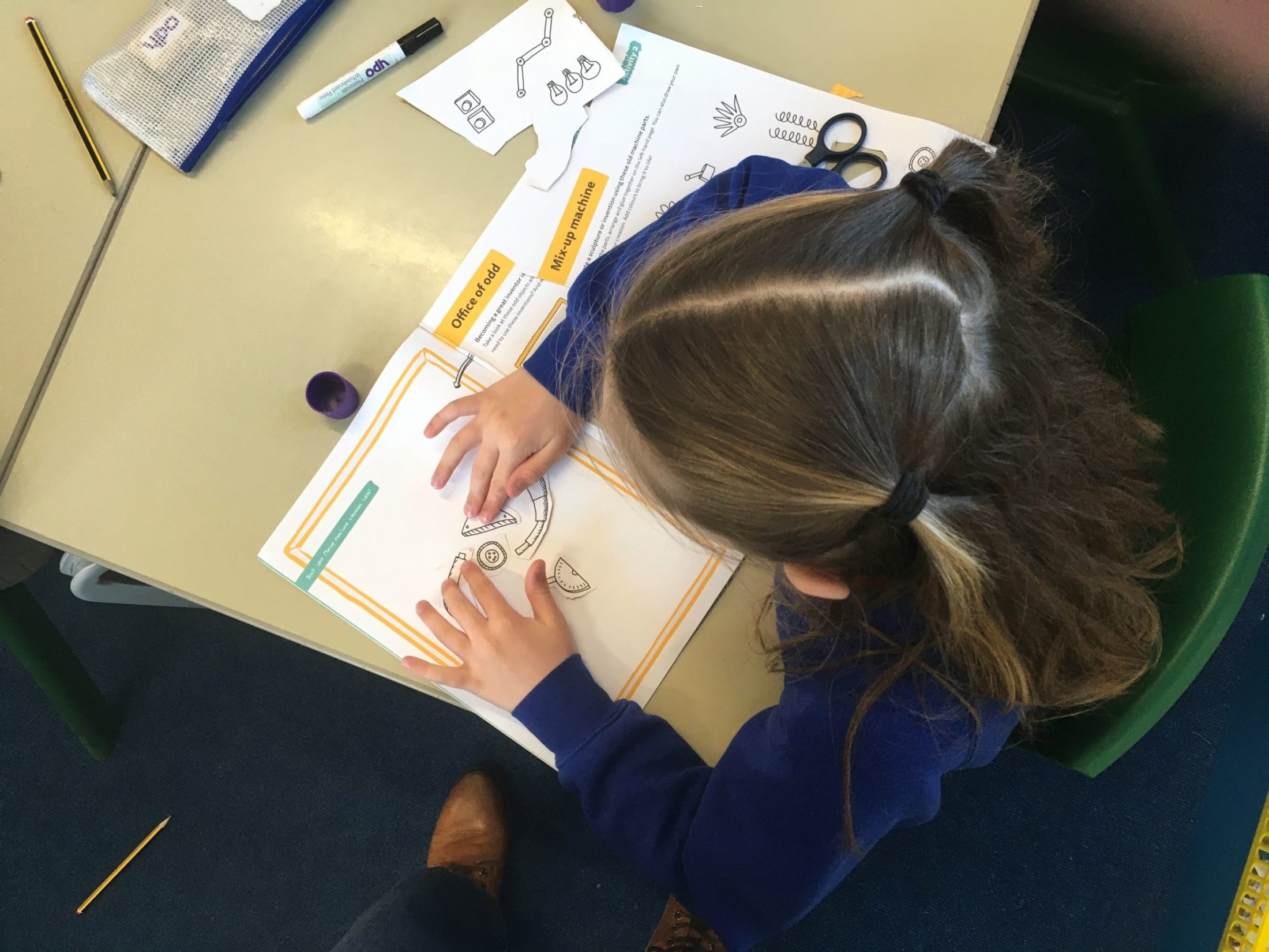 A young child with long brown hair sits at a desk, colouring and cutting out printed worksheets with diagrams and illustrations. Scissors, a glue stick, and other stationery items are on the table.
