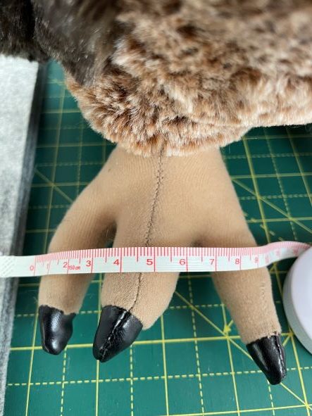 A plush animal's paw with black claws is being measured with a tape measure, showing a width of about 4 inches. The paw rests on a green cutting mat with a yellow grid.
