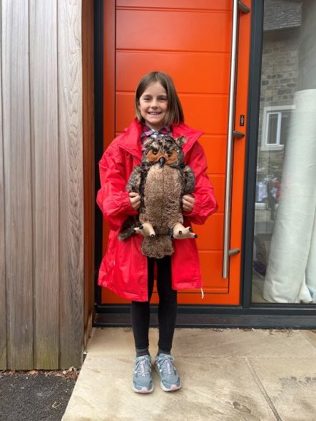 A smiling young girl in a bright red raincoat stands outside near an orange door, holding a large plush owl toy. She is wearing dark leggings and grey trainers.