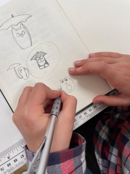 A person drawing owls with pencil in a dotted notebook; their left hand holds the page while the right hand sketches. A ruler and part of a checked shirt are visible.