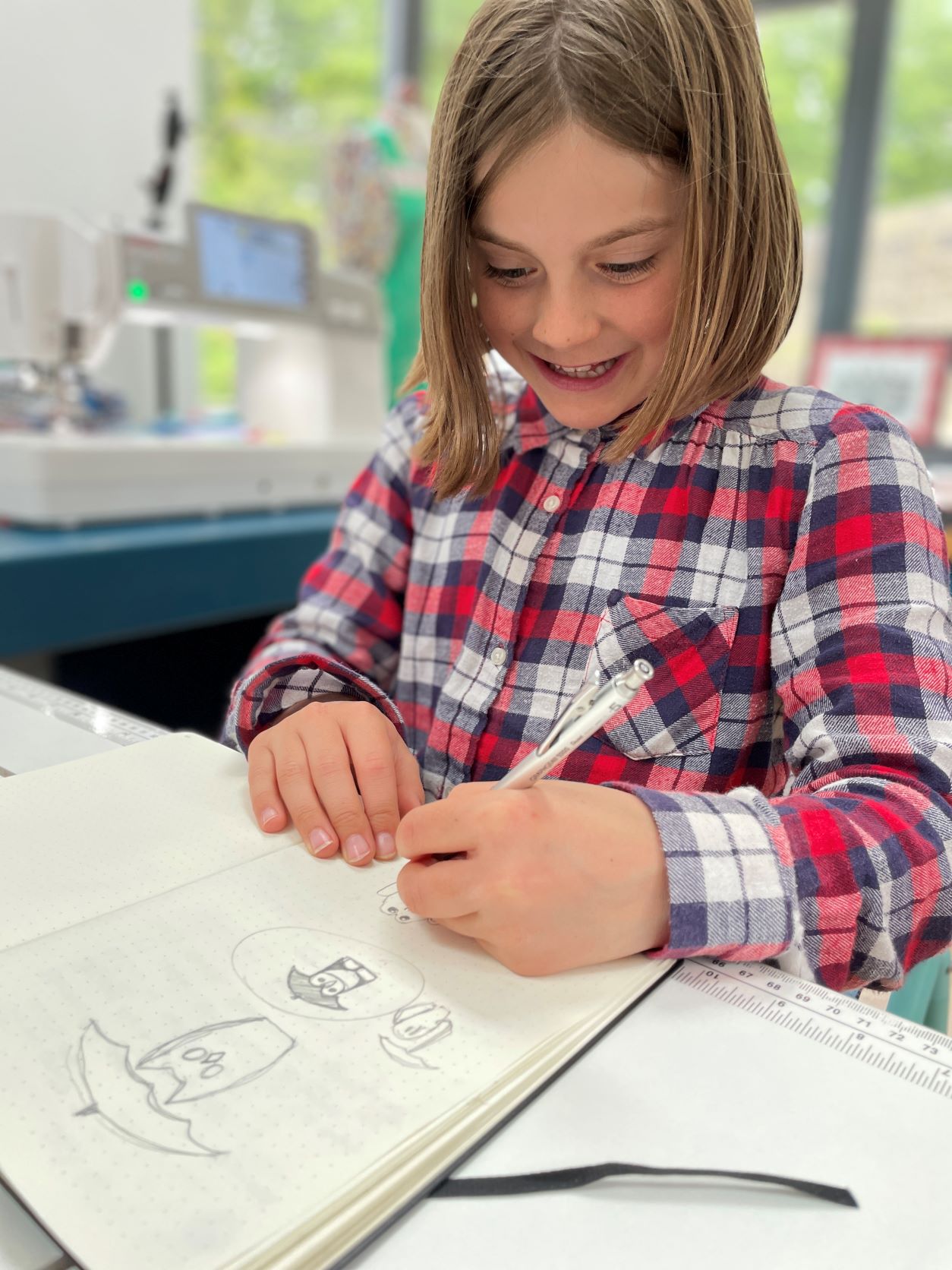 A smiling child in a red and blue checked shirt sketches cartoons in a notebook with a pencil, sitting at a bright desk with sewing tools and a ruler nearby.