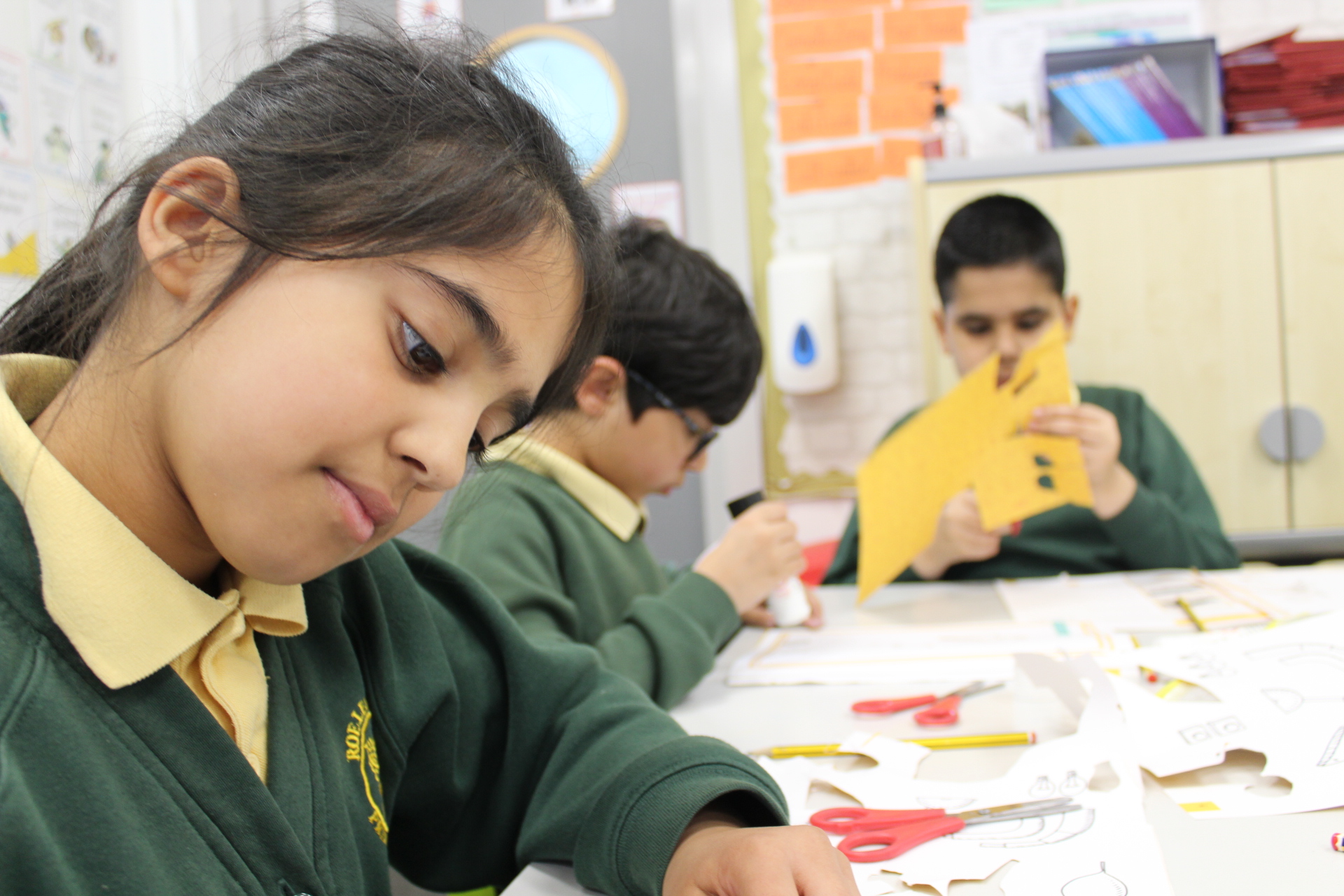 Three children in green school uniforms sit at a table doing arts and crafts. The girl in the foreground focuses on her project, while the two boys in the background cut and glue yellow paper. Classroom materials are scattered on the table.