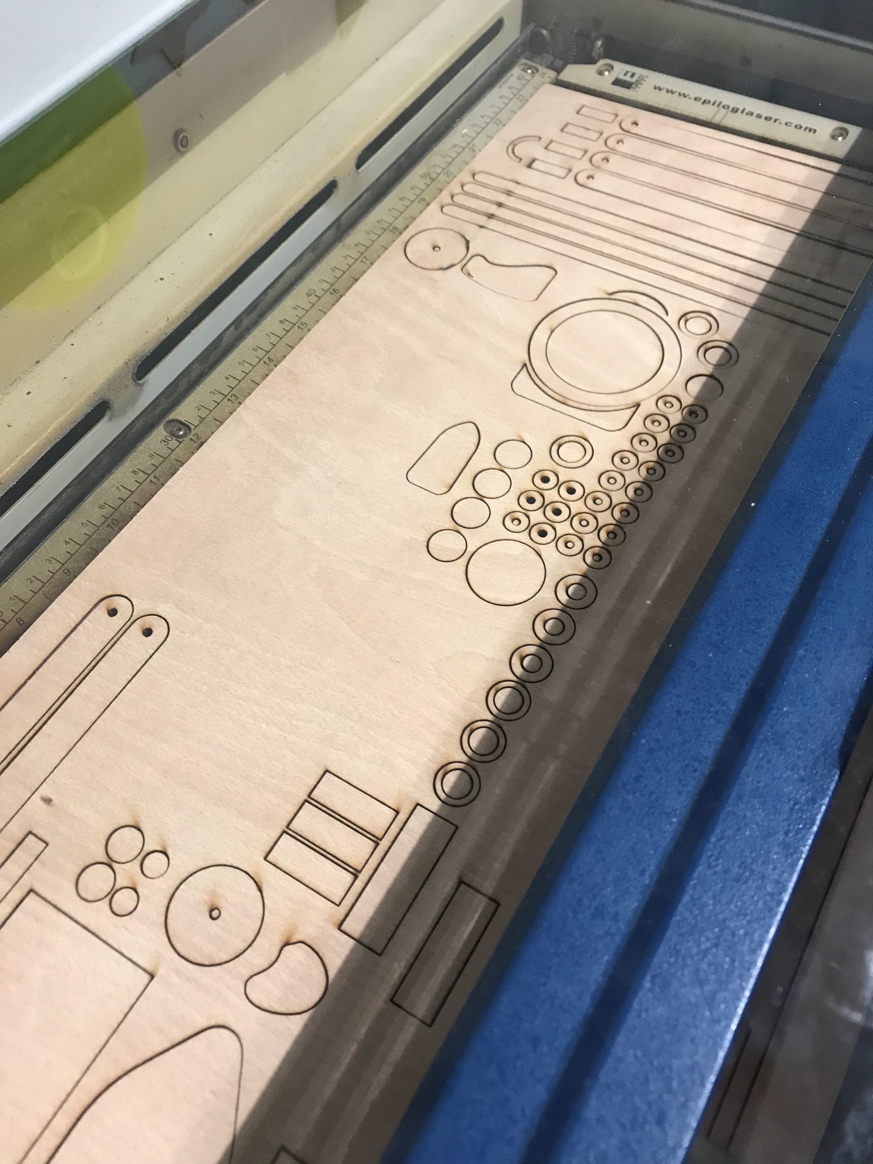 A laser cutting machine is engraving and cutting various geometric shapes and patterns into a rectangular sheet of light-coloured wood. The shapes include circles, rectangles, and ovals arranged in a neat layout.