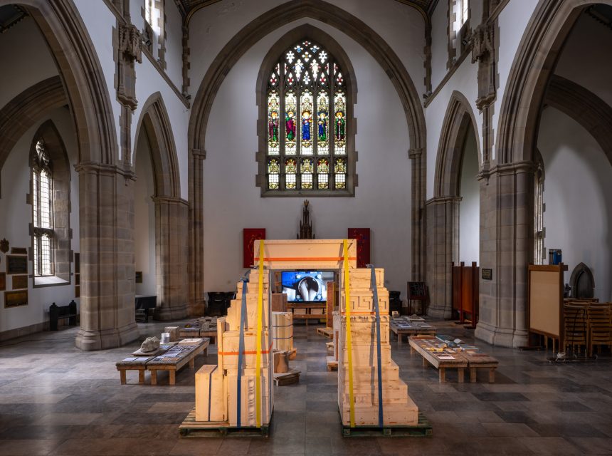 A large, ornate church interior with arched ceilings and stained glass windows, featuring a contemporary art installation of wooden crates and pallets arranged symmetrically down the nave.