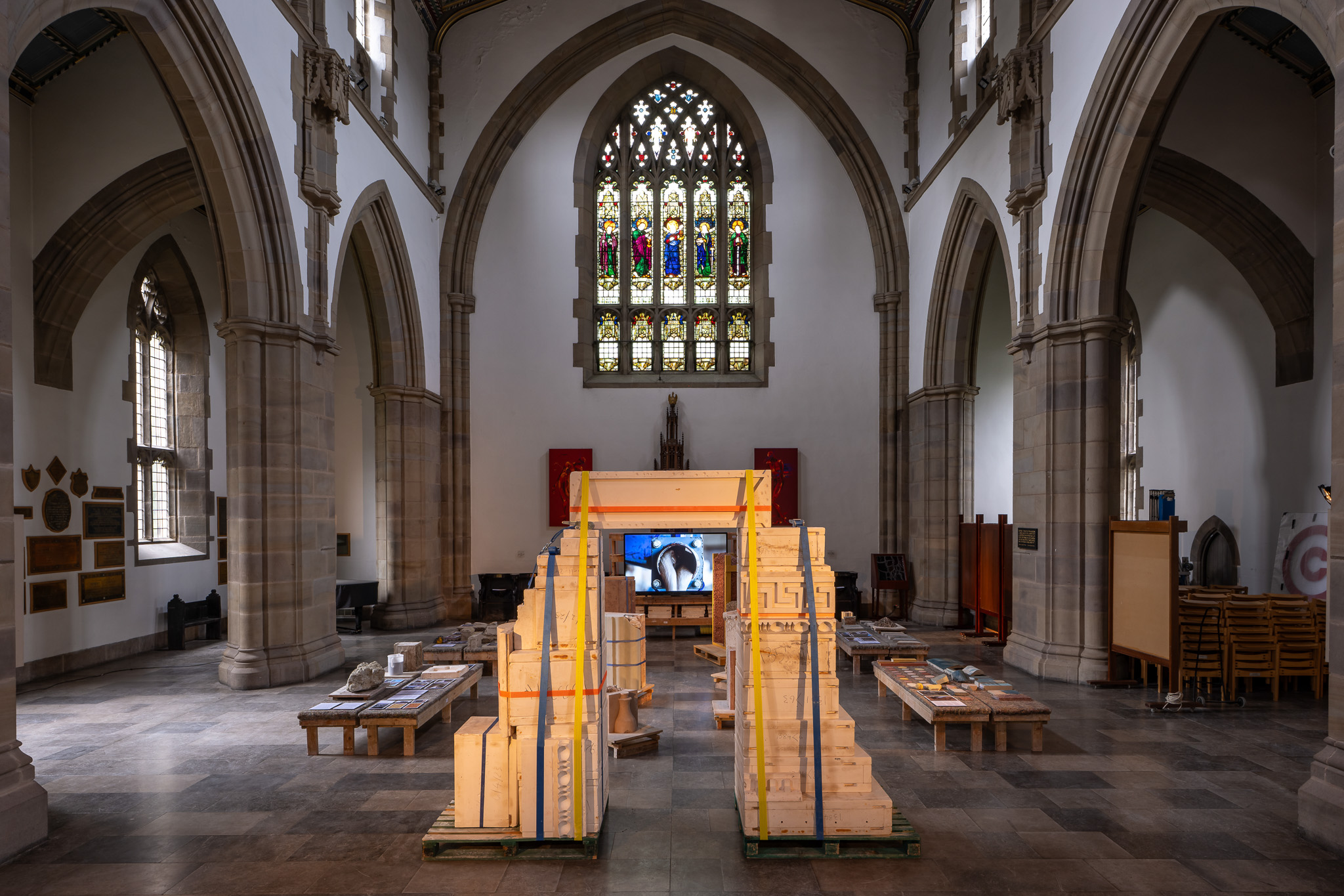 A large, ornate church interior with arched ceilings and stained glass windows, featuring a contemporary art installation of wooden crates and pallets arranged symmetrically down the nave.