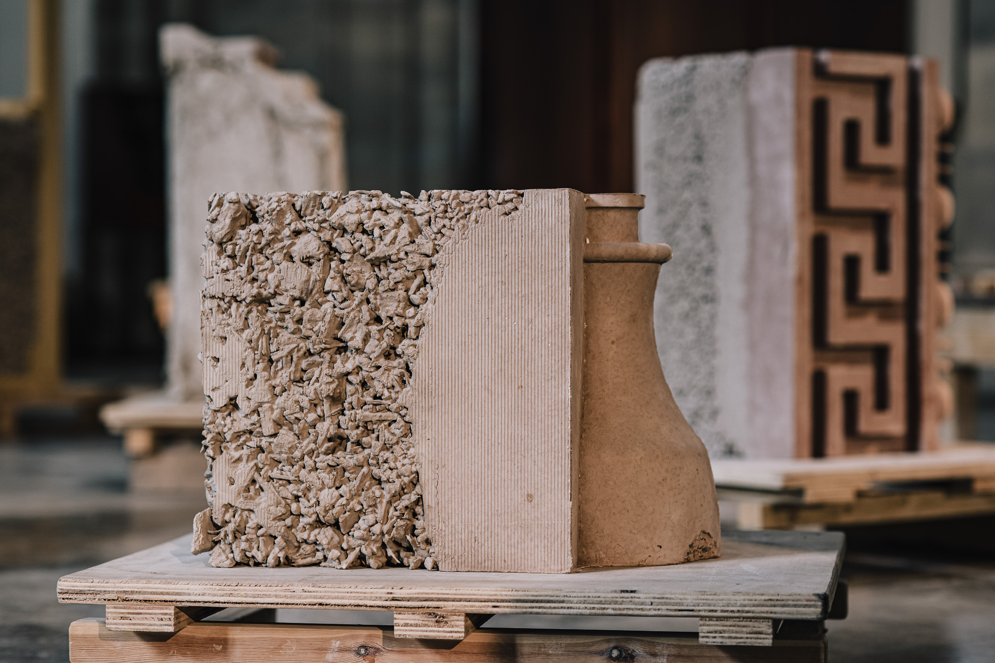 A sculptural artwork combining rough, textured fragments with smooth, curved ceramic surfaces, displayed on a wooden pallet in an indoor gallery setting. Other abstract pieces are visible in the background.