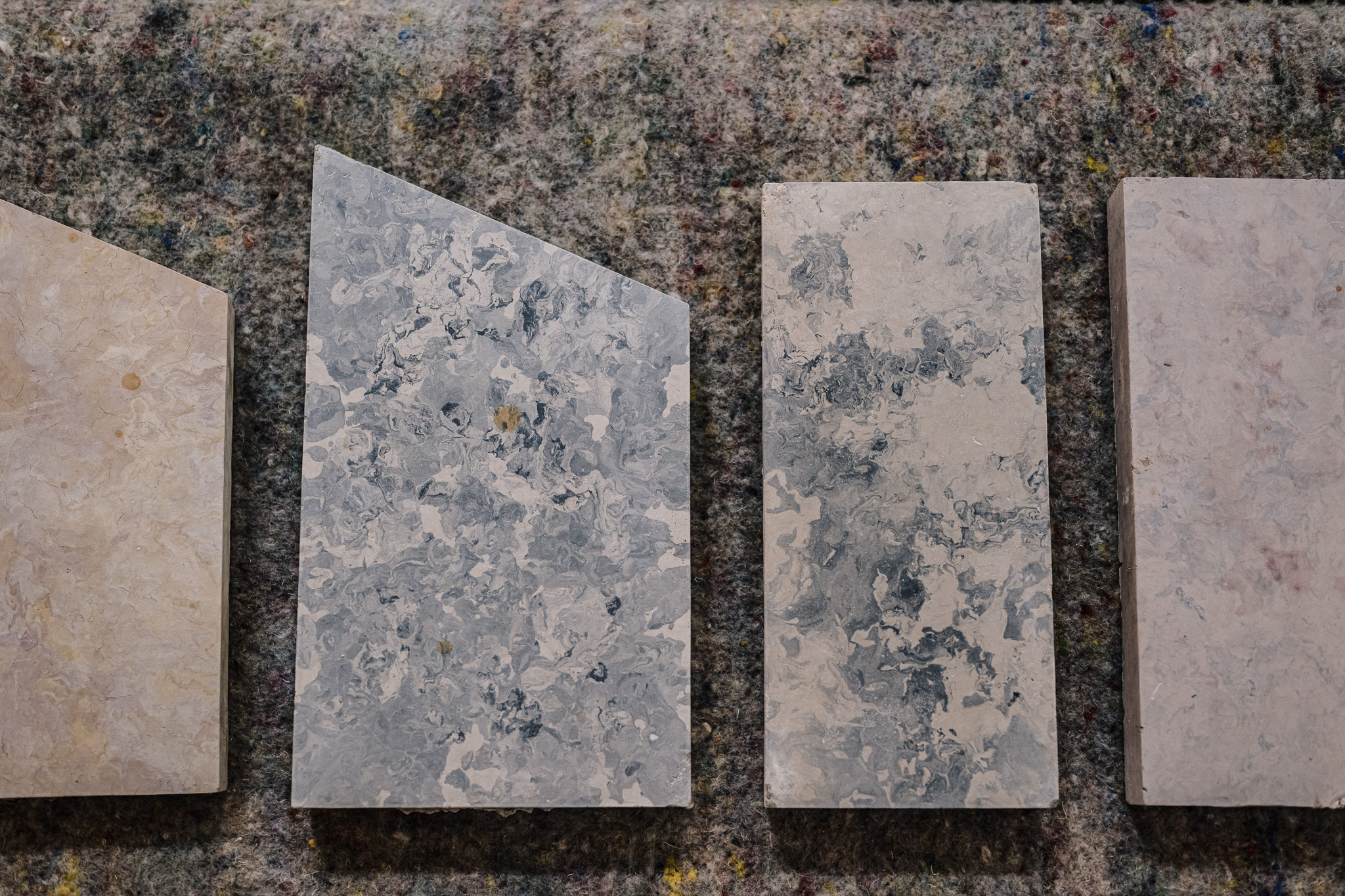 Four rectangular stone tiles with marbled grey, white, and beige patterns are arranged in a row on a textured, multicoloured surface. The second tile from the left has a diagonal top edge.