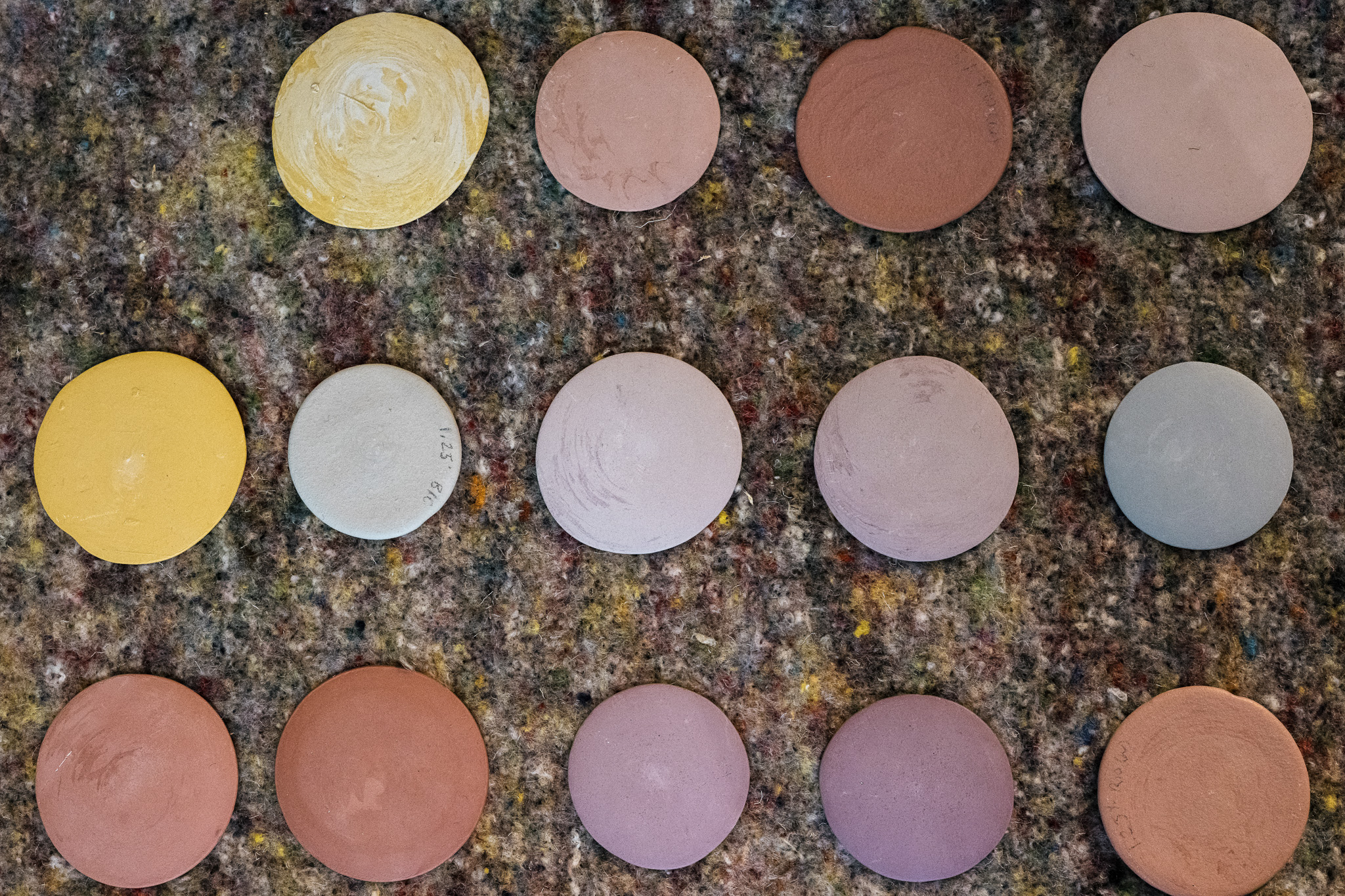Fifteen round, flat clay discs in various earthy colours are arranged in three rows on a speckled, textured surface. The discs vary in shades of yellow, brown, pink, and grey.