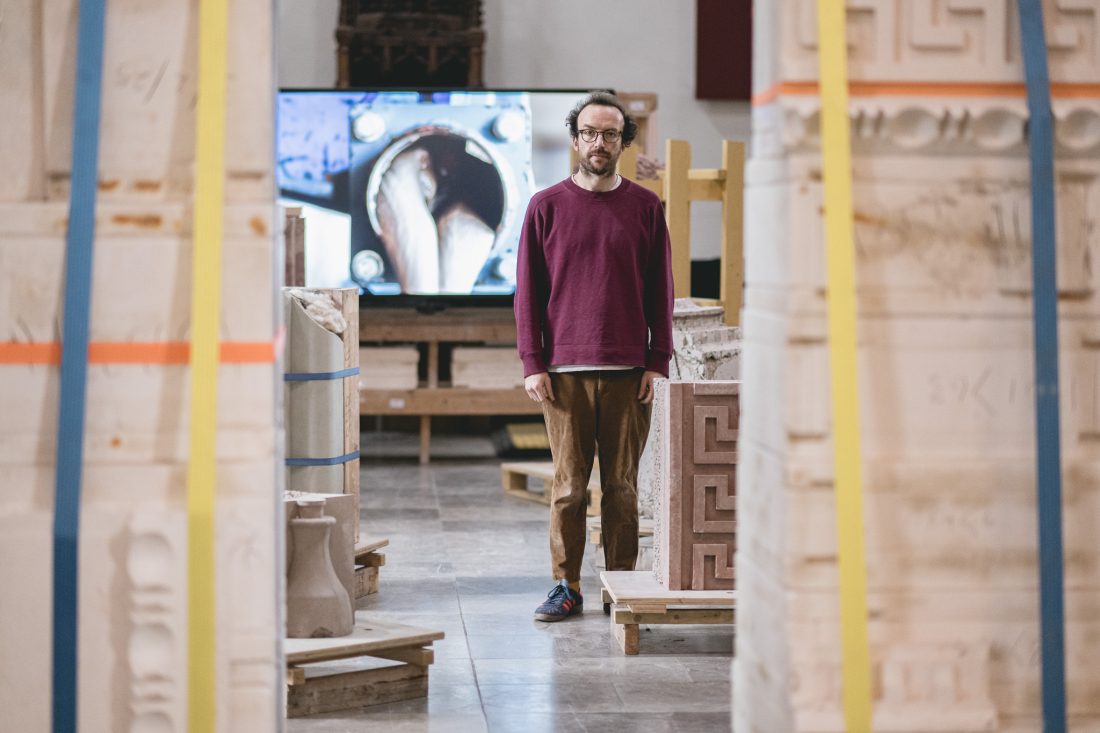 Lewis Jones in a maroon jumper and brown trousers stands among large sculpted blocks in an indoor space, with a screen showing a mechanical image behind him.