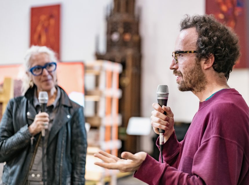 Two people holding microphones are engaged in conversation indoors. The man in the foreground gestures while speaking, and the woman in the background listens. Both wear glasses. Blurred artwork and shelves are visible behind them.