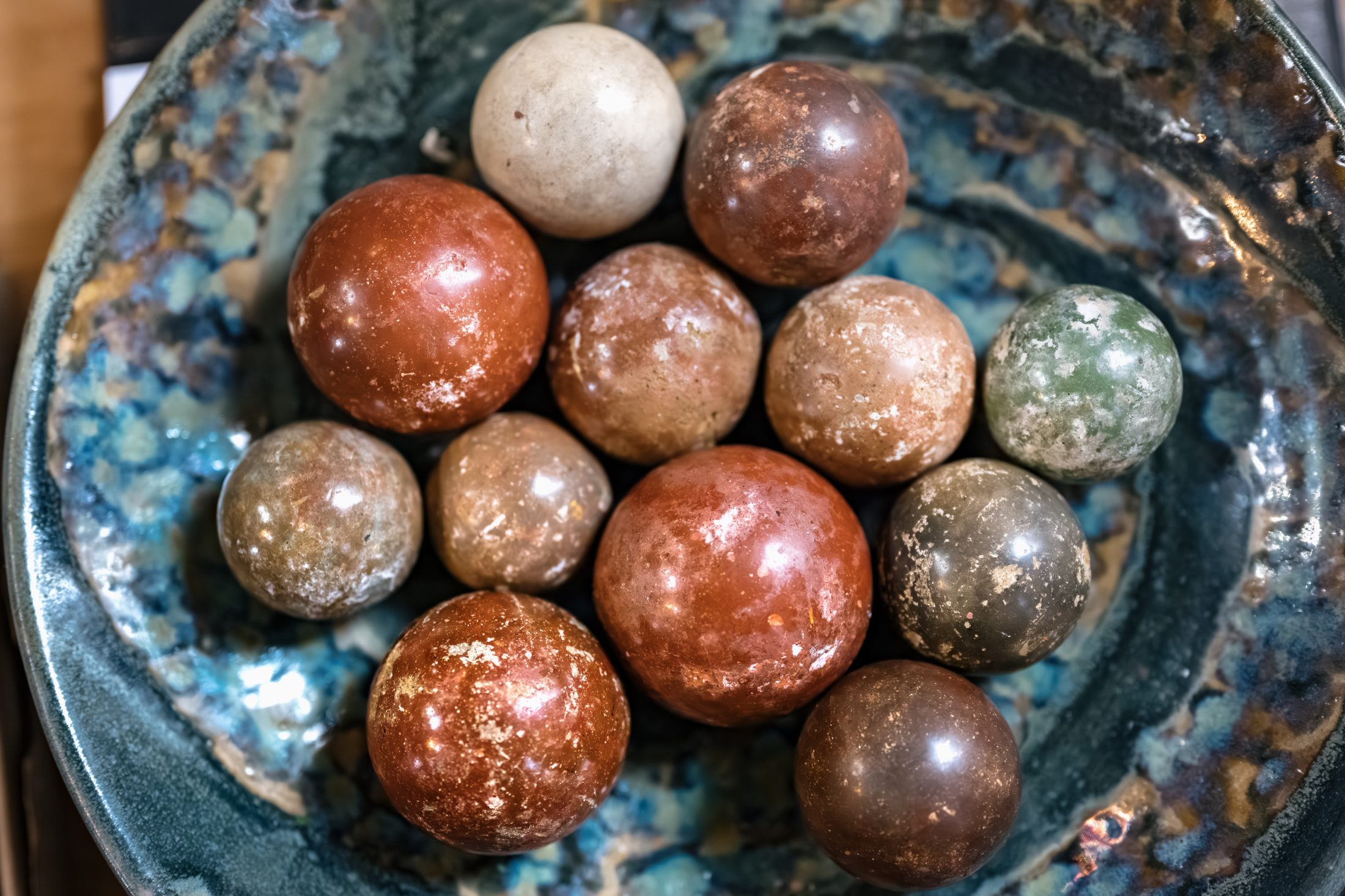 A ceramic bowl holds an assortment of round, polished stones in various natural colours including red, brown, tan, grey, and green, with a slightly weathered appearance.