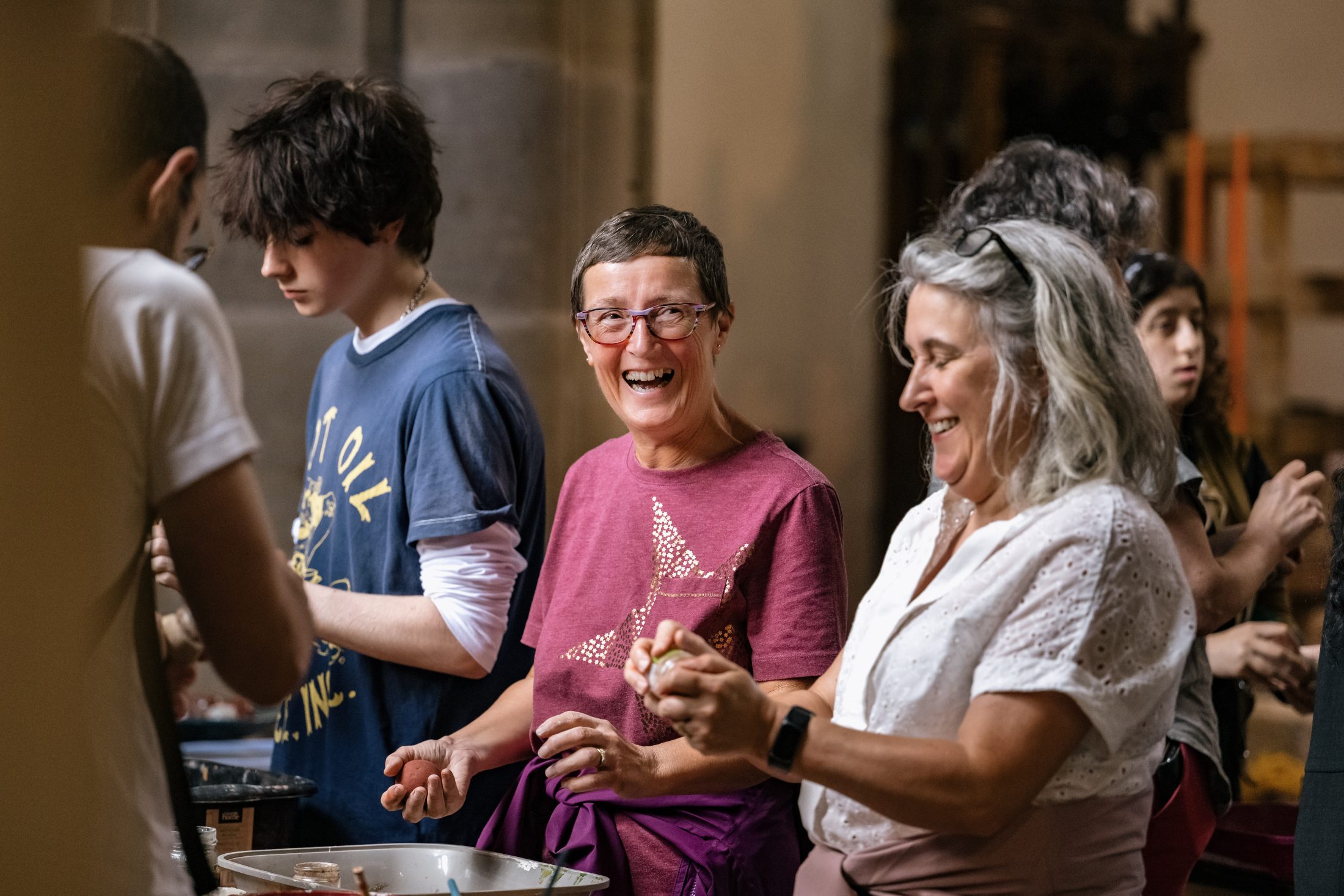 A group of people stand together preparing food, smiling and laughing. A woman in a purple shirt and glasses is at the centre, next to a woman with grey hair in a white blouse. The atmosphere is joyful and friendly.