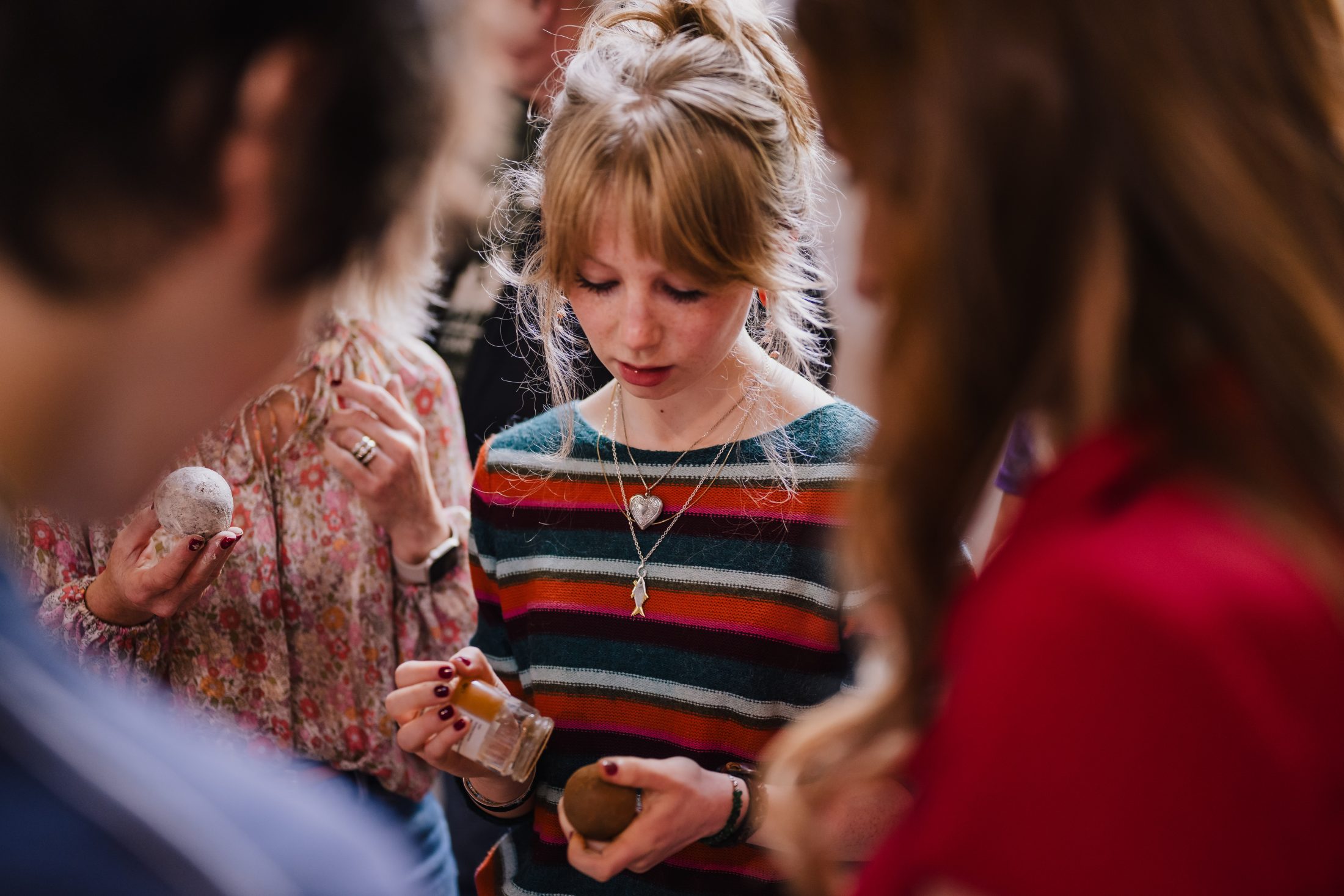 A young person in a striped jumper examines an object in their hands, surrounded by others who are also holding small items. The focus is on the person in the centre, with the others slightly out of focus.