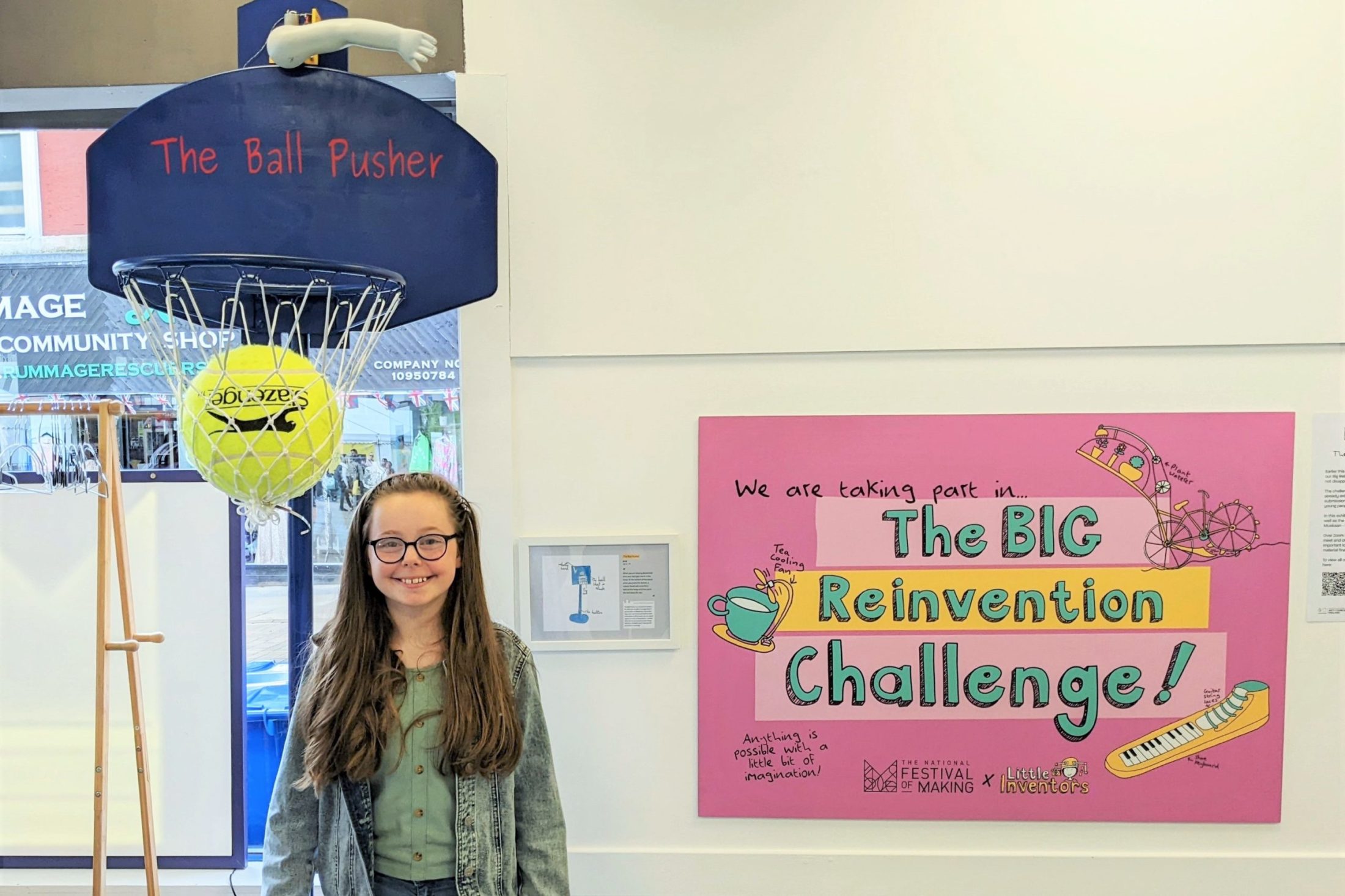 A girl with long hair and glasses stands smiling beside a pink sign that reads “The BIG Reinvention Challenge!” A large yellow tennis ball sits in a basketball net above her, labelled The Ball Pusher.